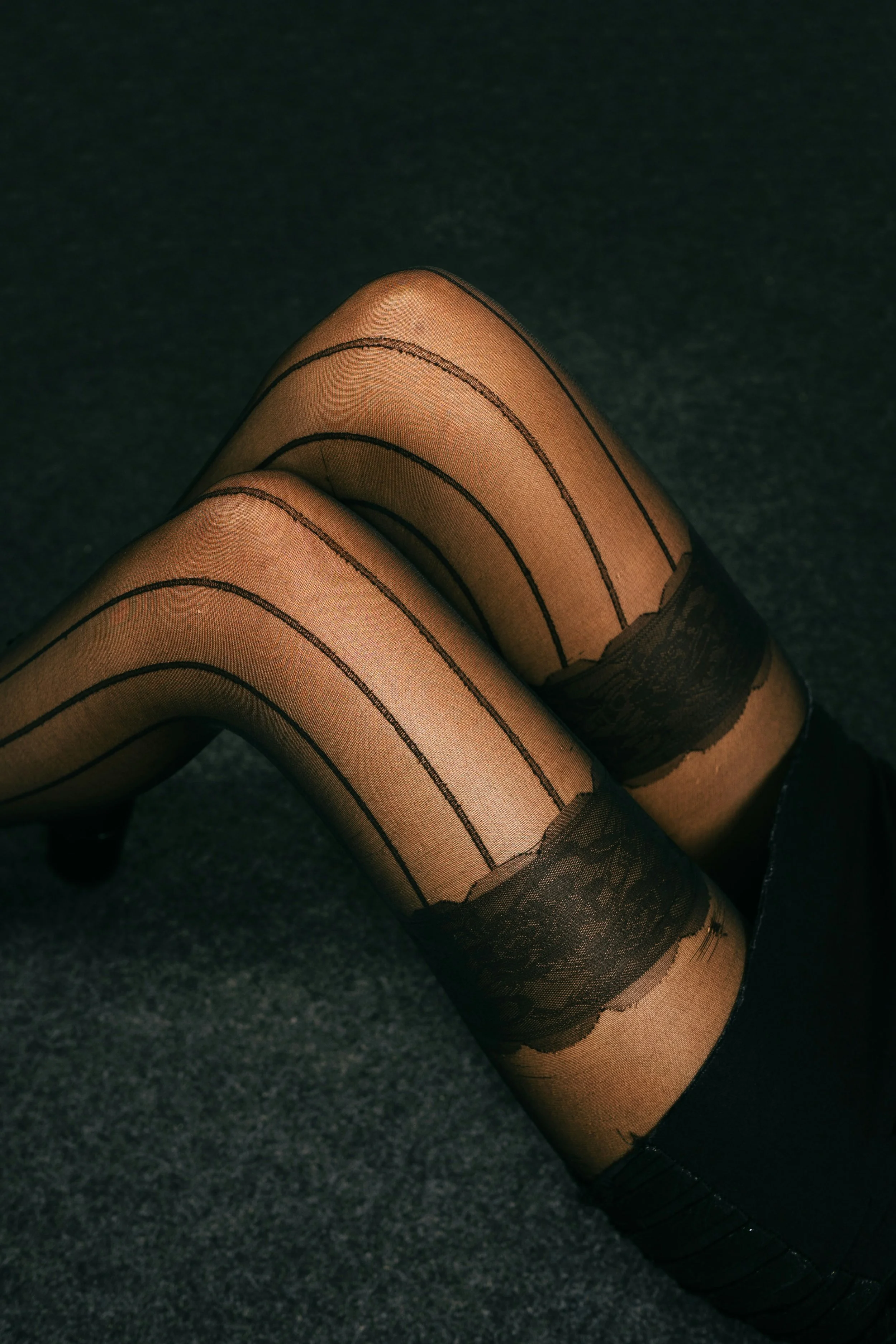 Close-up of a person's legs wearing patterned black stockings with horizontal black lines, sitting on a dark surface, with the black shoes partially visible.