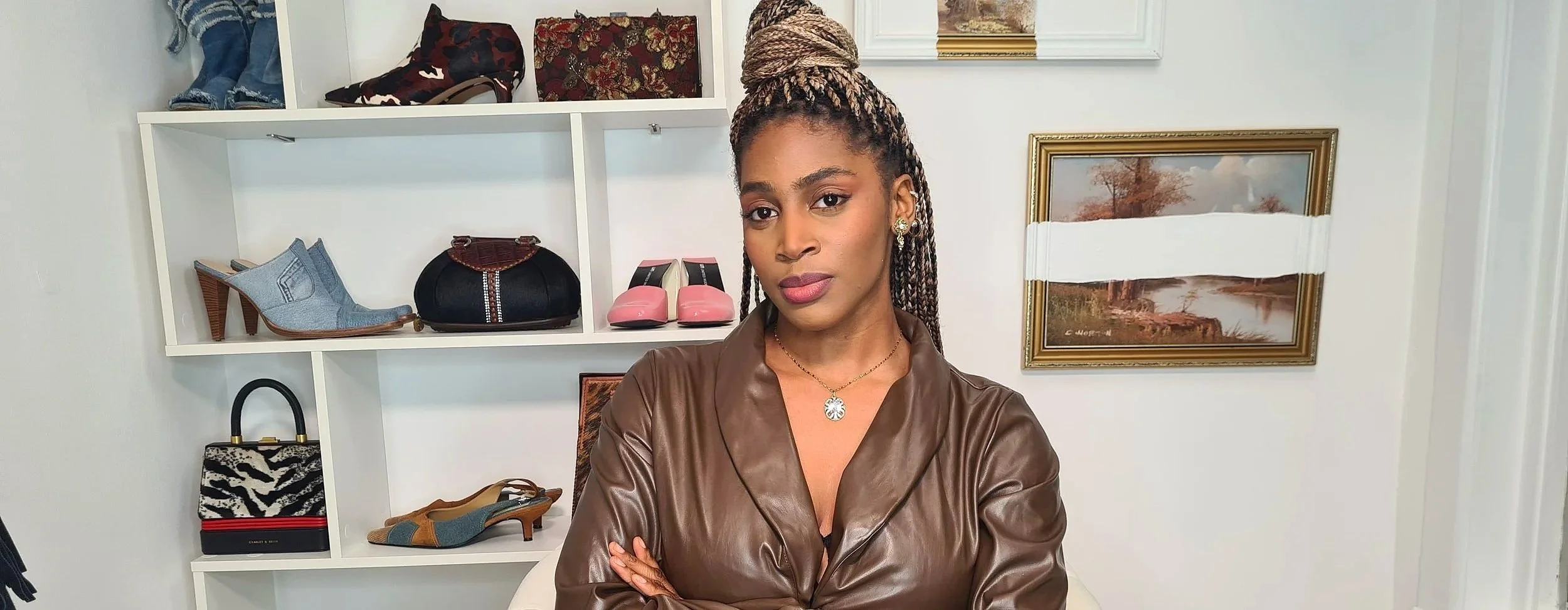 A woman with braided hair wearing a brown leather jacket and gold jewelry, standing in front of a white shelving unit filled with various shoes, handbags, and accessories, and a landscape painting on the wall.