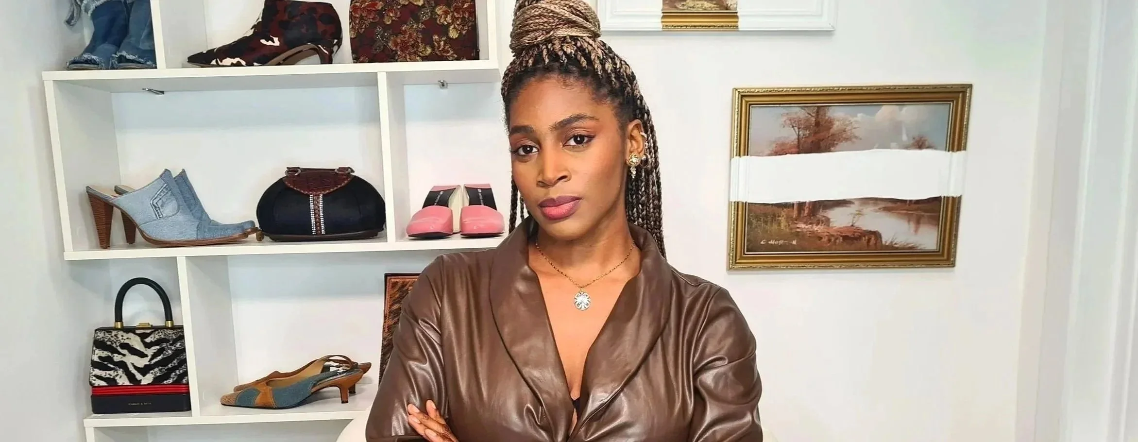 A woman with braided hair in a bun, wearing a brown leather jacket and jewelry, standing in front of a white shelf with shoes, handbags, and decor items, and a framed landscape painting on the wall.