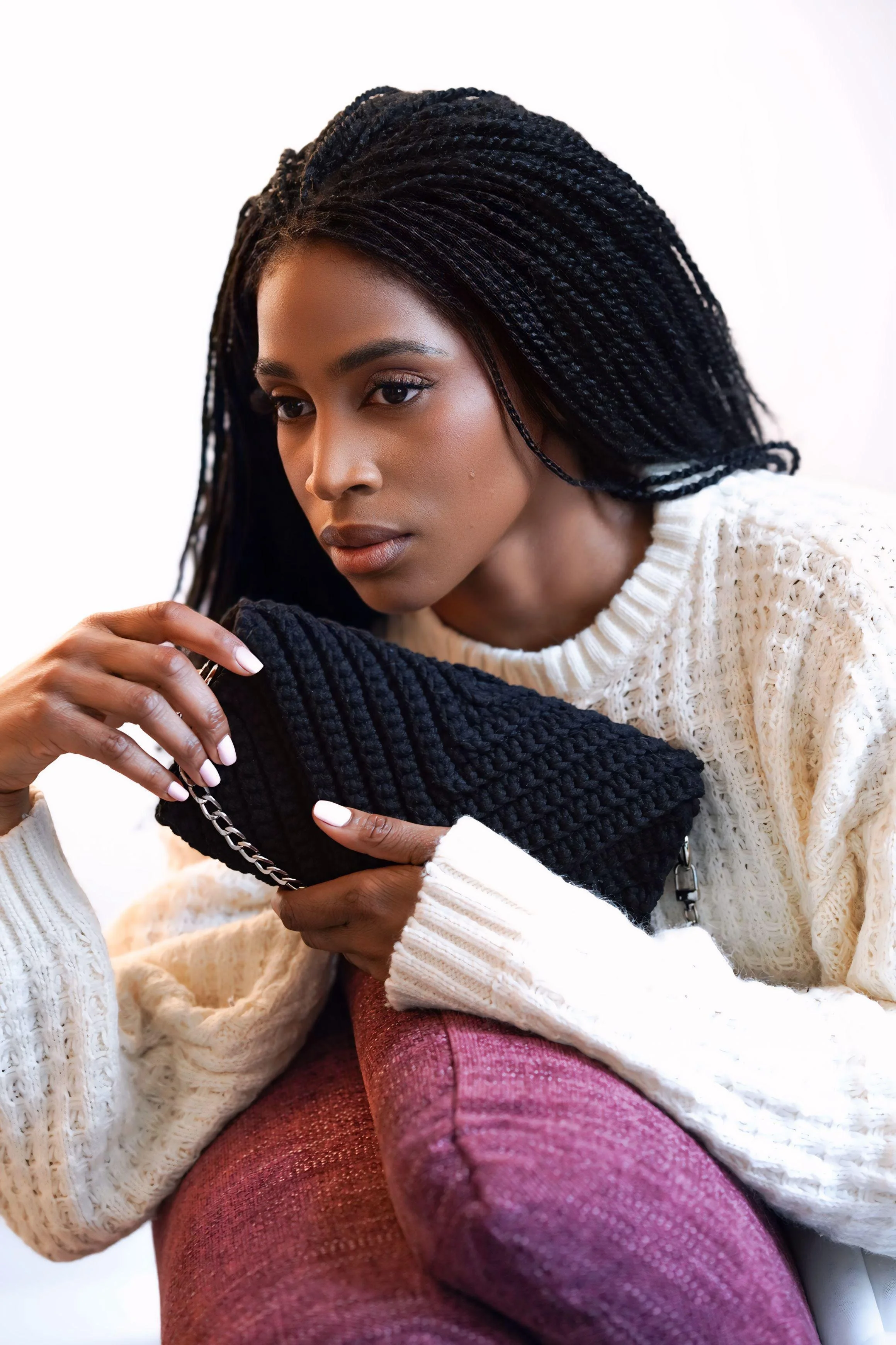A woman with long, twisted black hair resting her head on her arm, holding a black purse with a chain strap, and wearing a white knit sweater and pink pants, against a plain background.