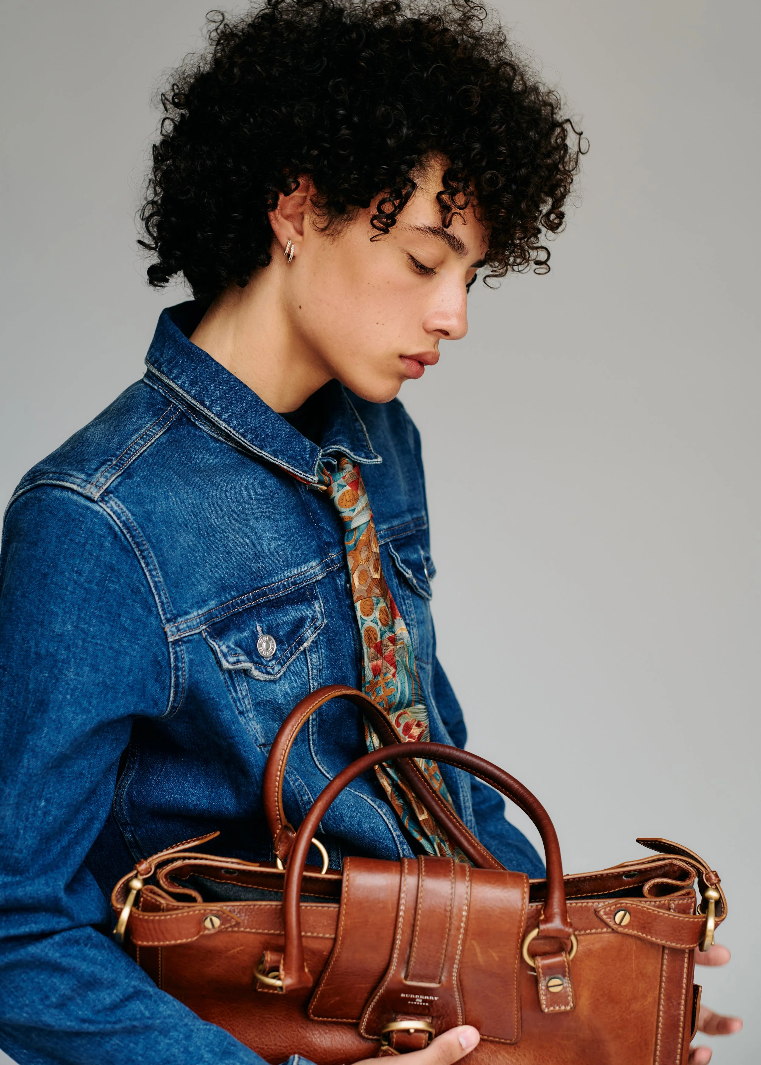 A woman with curly black hair, wearing a denim jacket, holding a brown leather handbag with gold hardware, standing against a plain gray background.