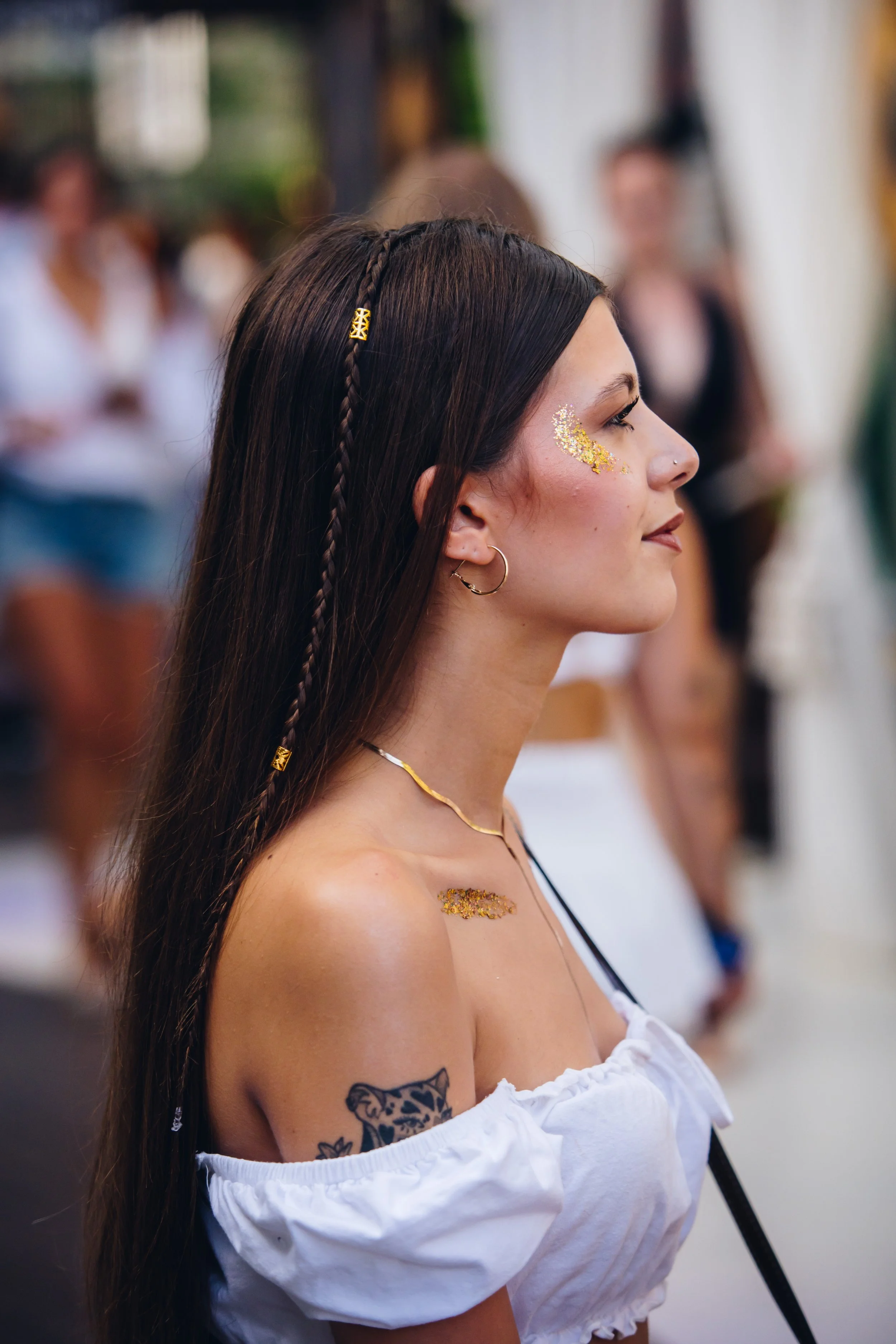 A young woman with long dark hair, braided and decorated with gold beads, smiling in profile. She has glitter on her cheek, wears hoop earrings, and a white off-the-shoulder top with a tattoo on her shoulder. The background shows a blurred group of people at an indoor event.