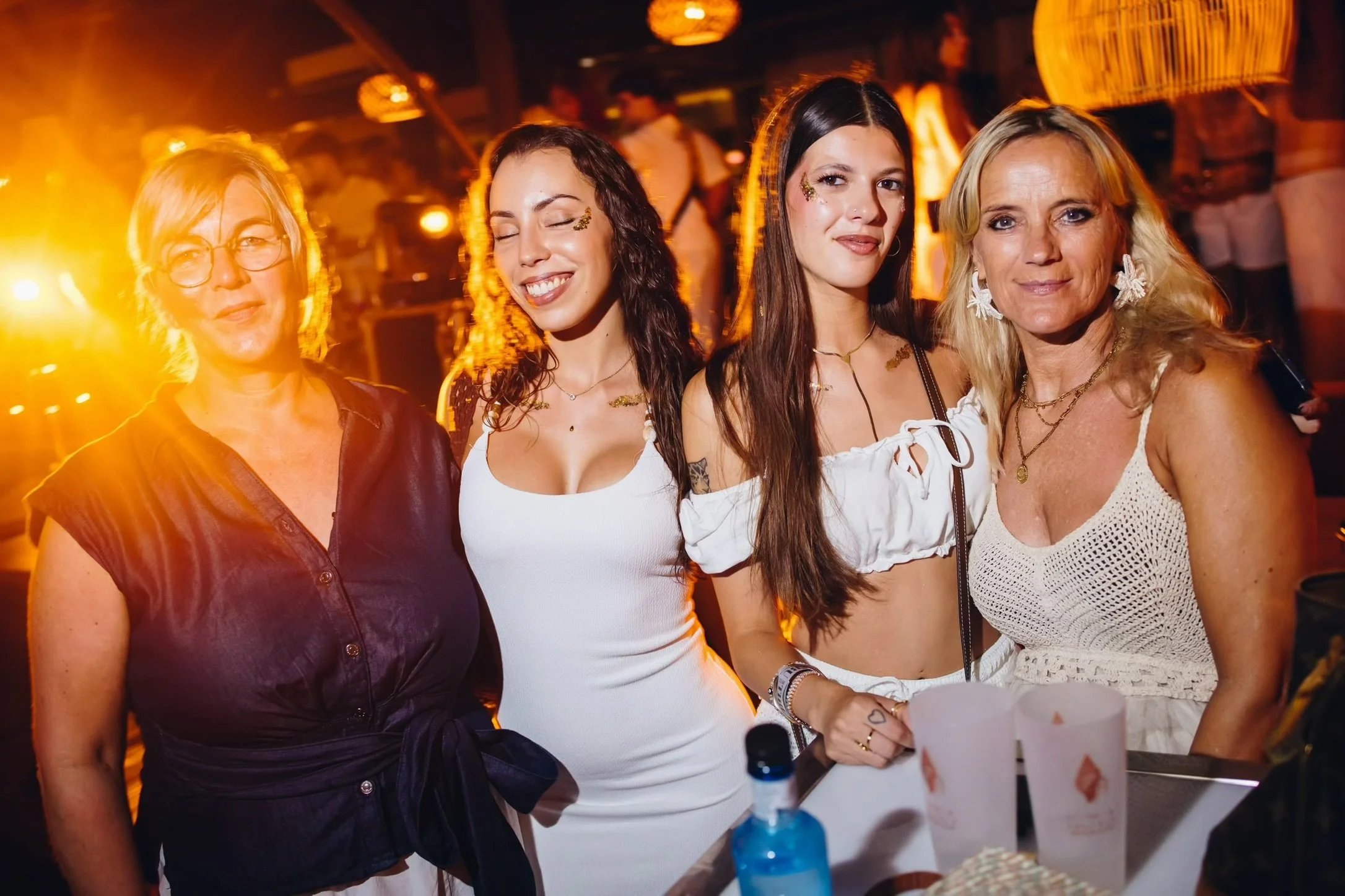 Four women at a party or celebration, standing close together, with bright yellow and orange lighting in the background.
