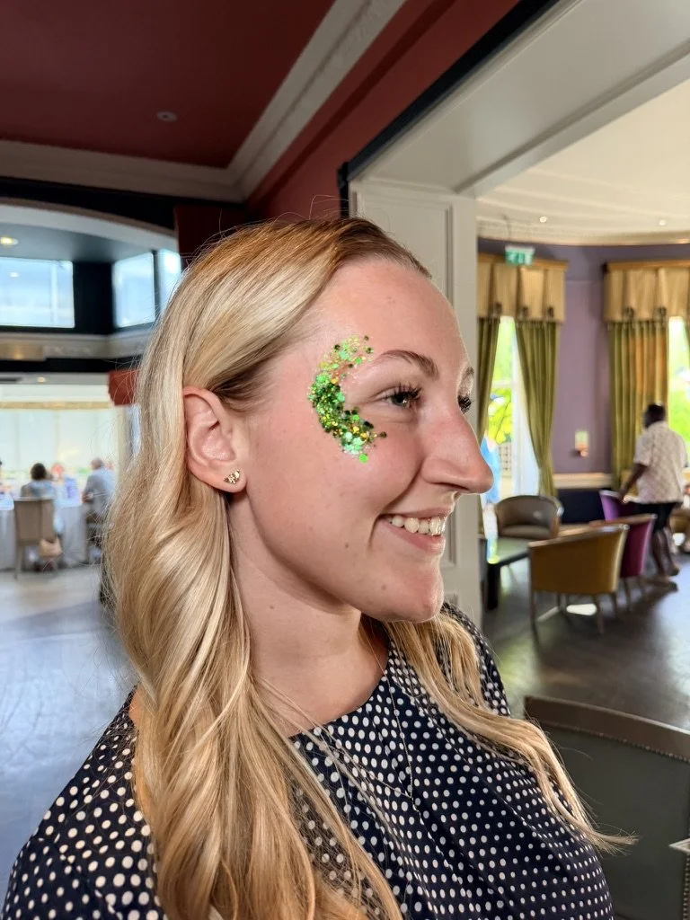 A young woman with long blonde hair, wearing a black and white polka dot top, smiling and showing green glitter and sequins on her right cheek in a brightly lit indoor setting.