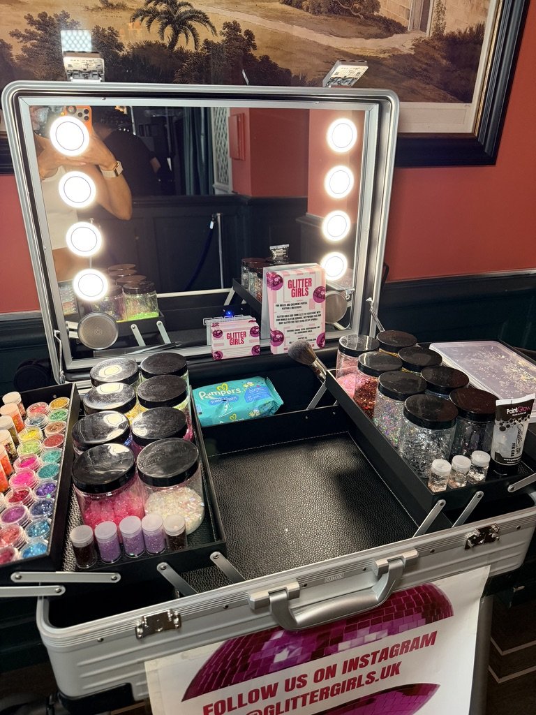 Travel-sized makeup and glitter supplies on a portable case with a mirror and ring light, advertising Glitter Girls.