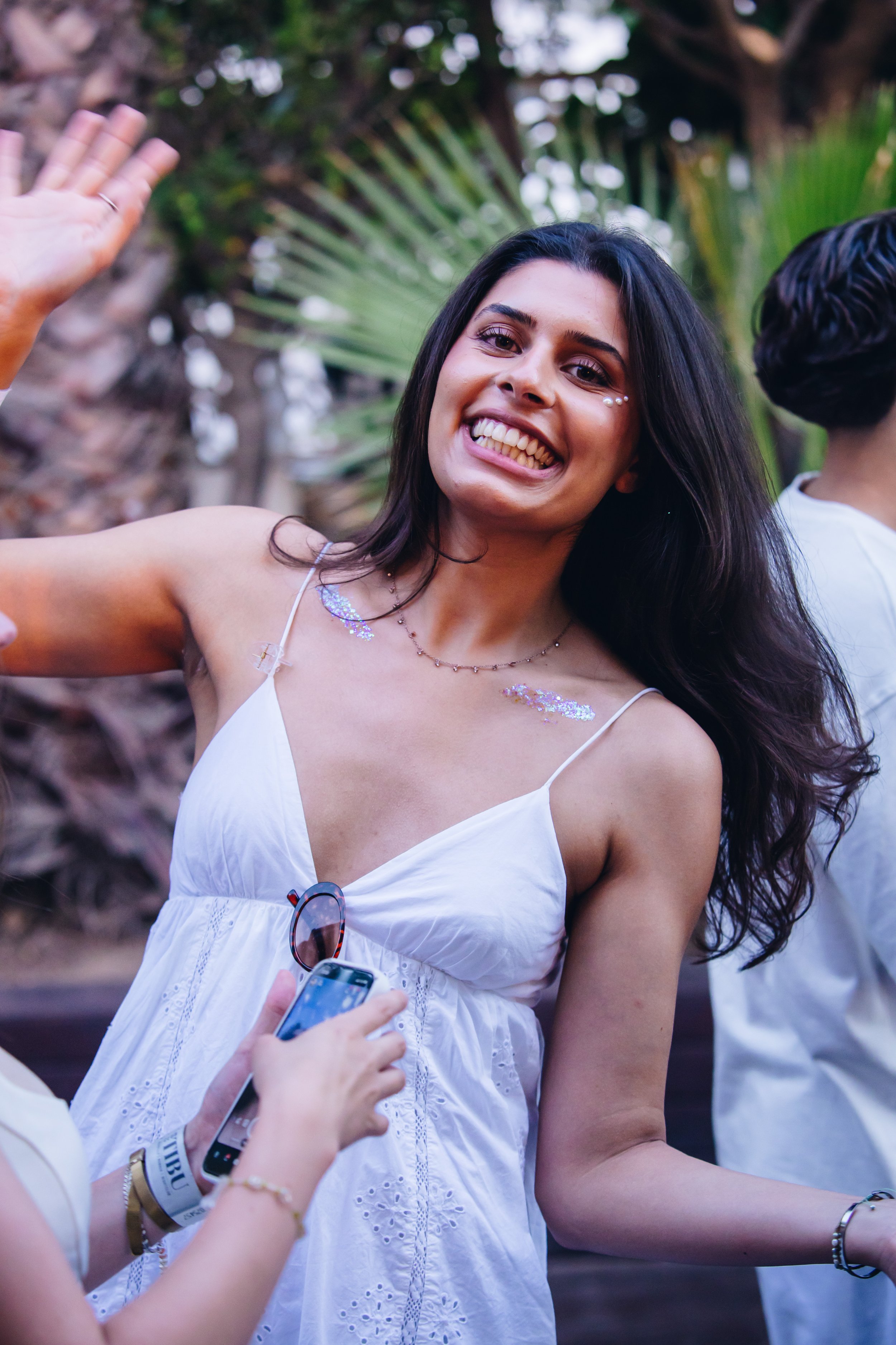 A woman with long dark hair smiling and waving at a social gathering in an outdoor setting with tropical plants in the background. She is wearing a white sleeveless dress with embroidery, small face decorations, and jewelry.