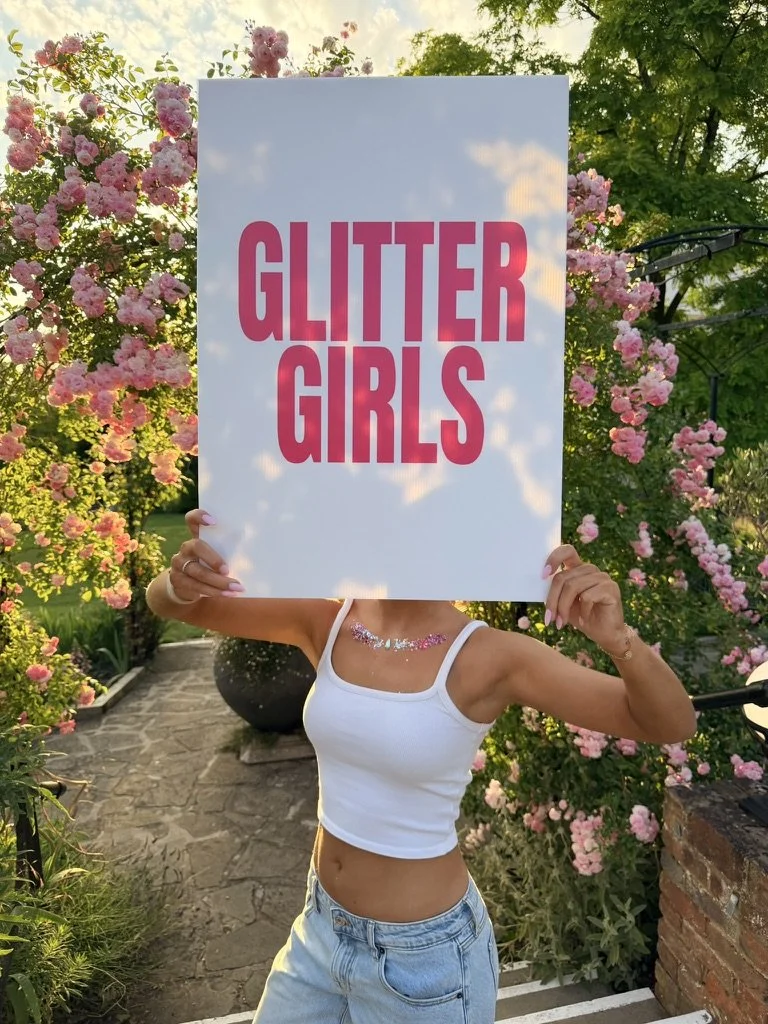 Woman holding a sign that reads 'Glitter Girls' in a garden with pink flowers and greenery