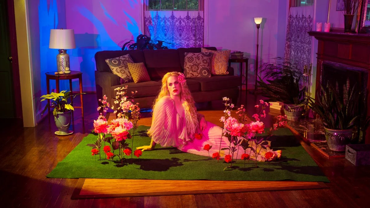 A woman with long, wavy blonde hair and pink flowers in her hair is sitting on a green rug decorated with artificial flowers in a living room. The room is illuminated with purple and pink lighting, with a dark sofa, various cushions, and plants in th