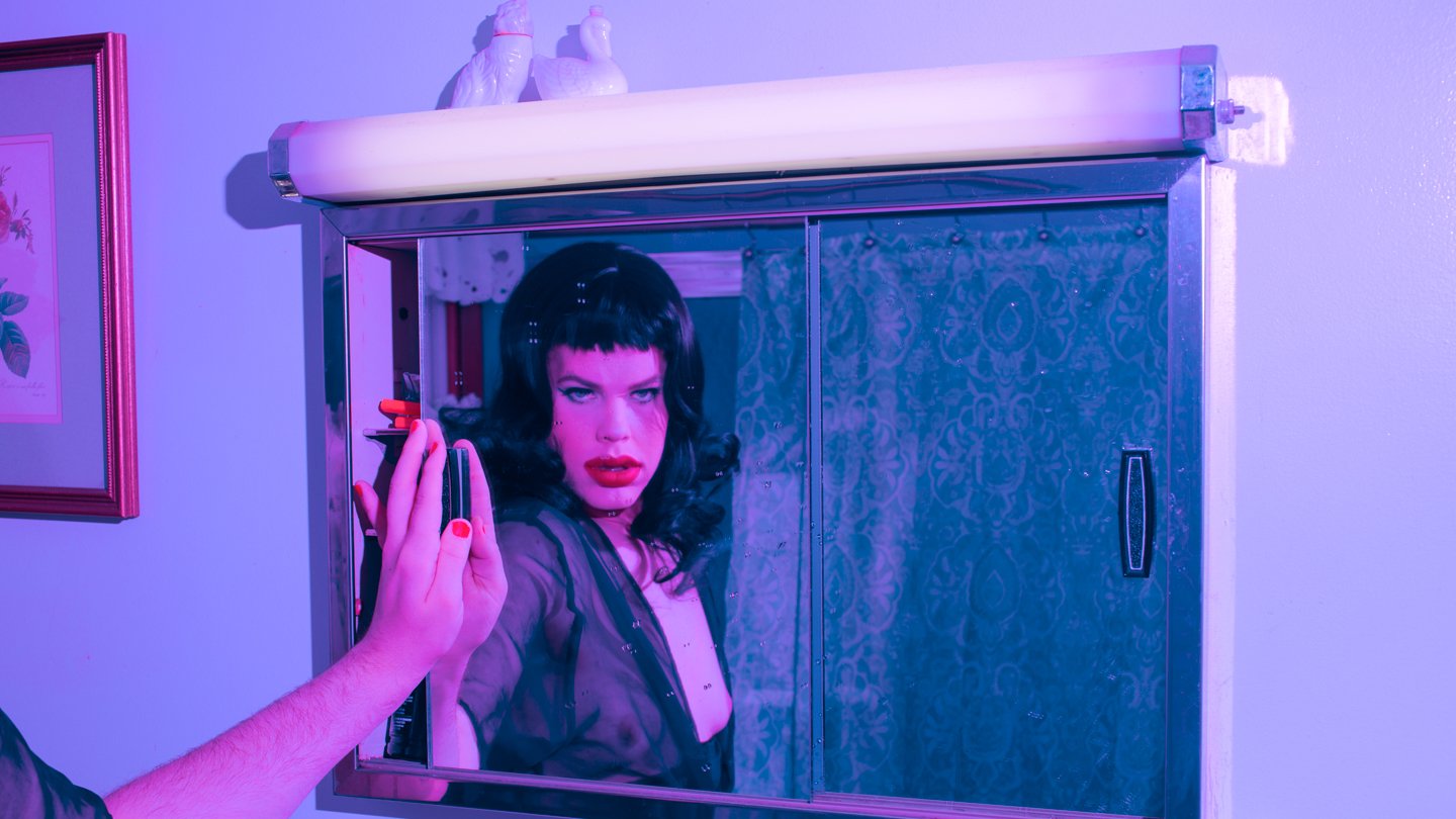 A woman with dark hair and red lipstick looking into a mirror, with a person's hand holding a makeup brush near her face; the scene is bathed in purple lighting.