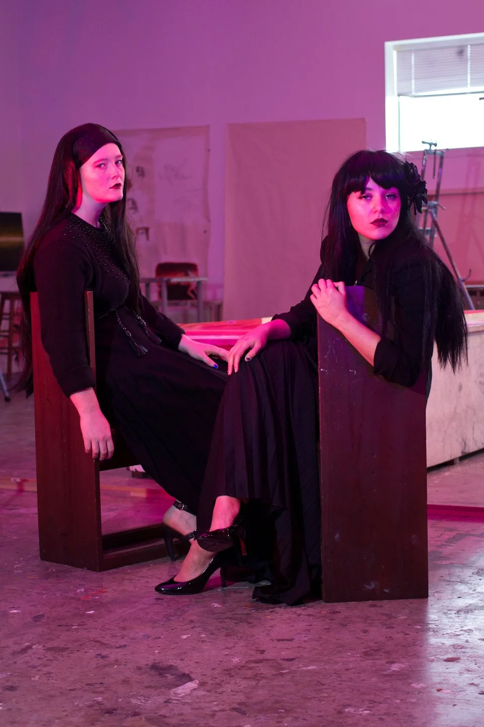 Two women with dark hair and gothic makeup sitting on wooden benches in a room with purple and pink lighting.
