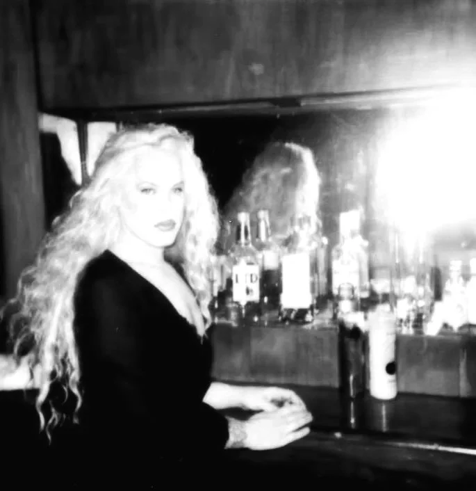 A woman with long, curly hair sitting at a bar with bottles of alcohol behind her and a mirror reflecting her image.