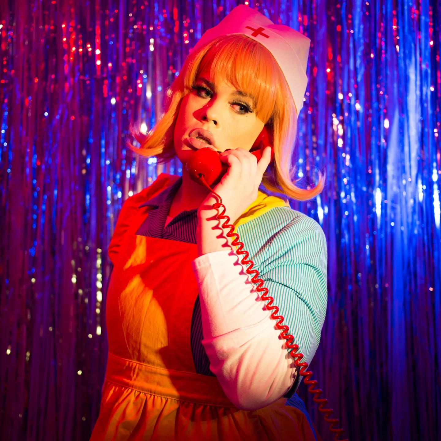 A woman dressed in a colorful nurse costume with a white nurse cap holding a red vintage telephone against a background of shiny blue and purple streamers