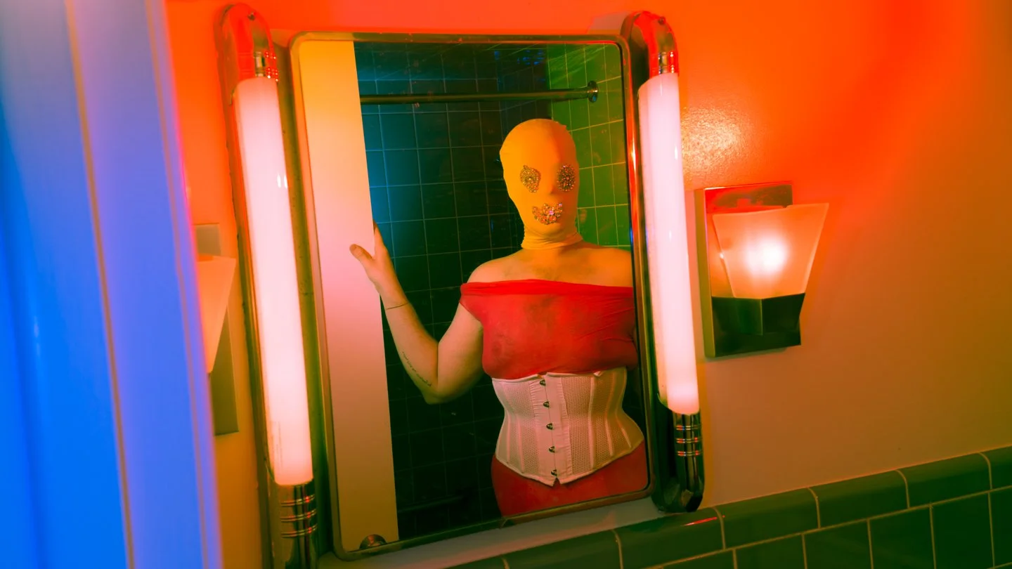 Person wearing a yellow mask with glittery decorations, a red off-the-shoulder top, a white corset, and a red skirt, standing in front of a mirror in a colorful, vibrant bathroom.