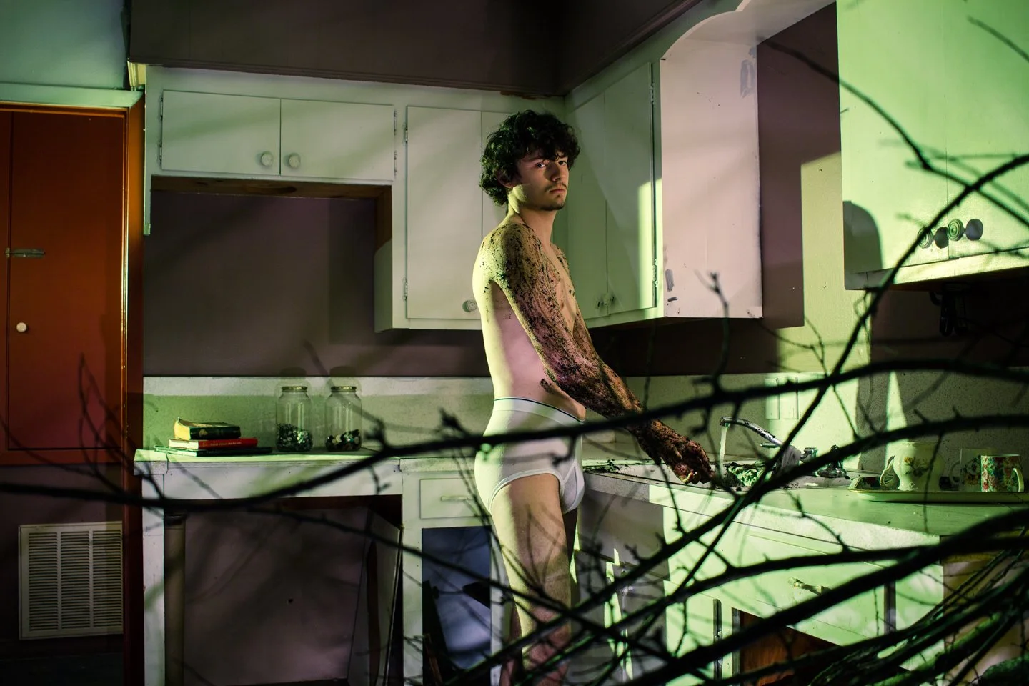 A young man with curly hair standing shirtless in a cluttered kitchen, covered in dirt or mud, wearing white shorts, with a serious expression, surrounded by shadows and dim lighting.