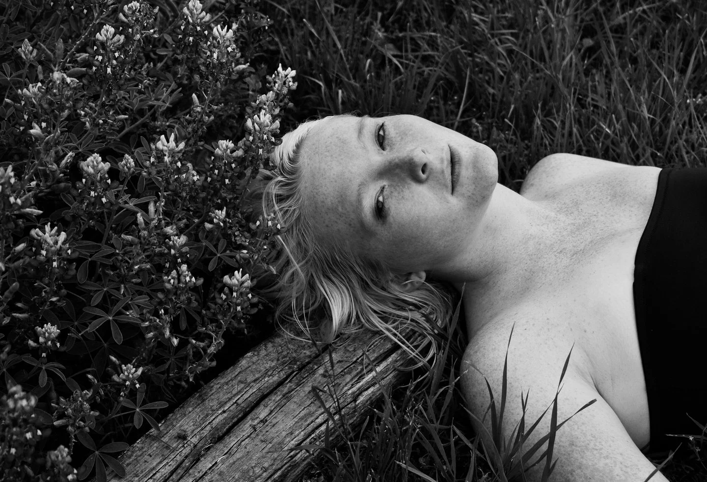 A black and white photo of a young woman with freckles lying on the grass next to a wooden log, with her head resting on a cluster of bushes, looking at the camera.