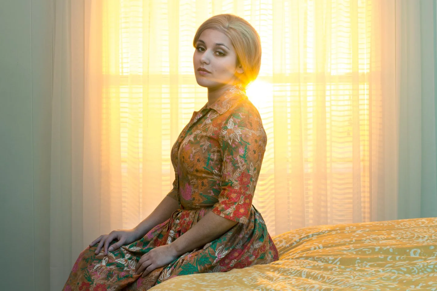 A woman with short blonde hair wearing a colorful floral dress, sitting on a bed, backlit by bright sunlight coming through a window with sheer curtains.