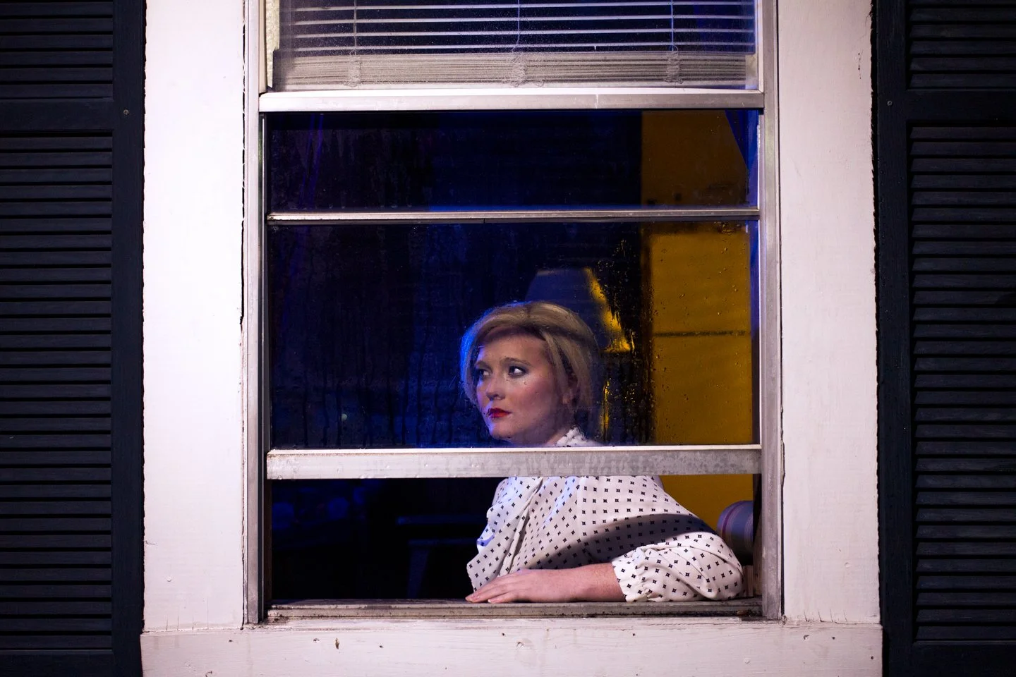 A woman with short blonde hair and red lipstick gazes out a window at night, wearing a white polka-dot blouse, with the window frame and black shutters visible.
