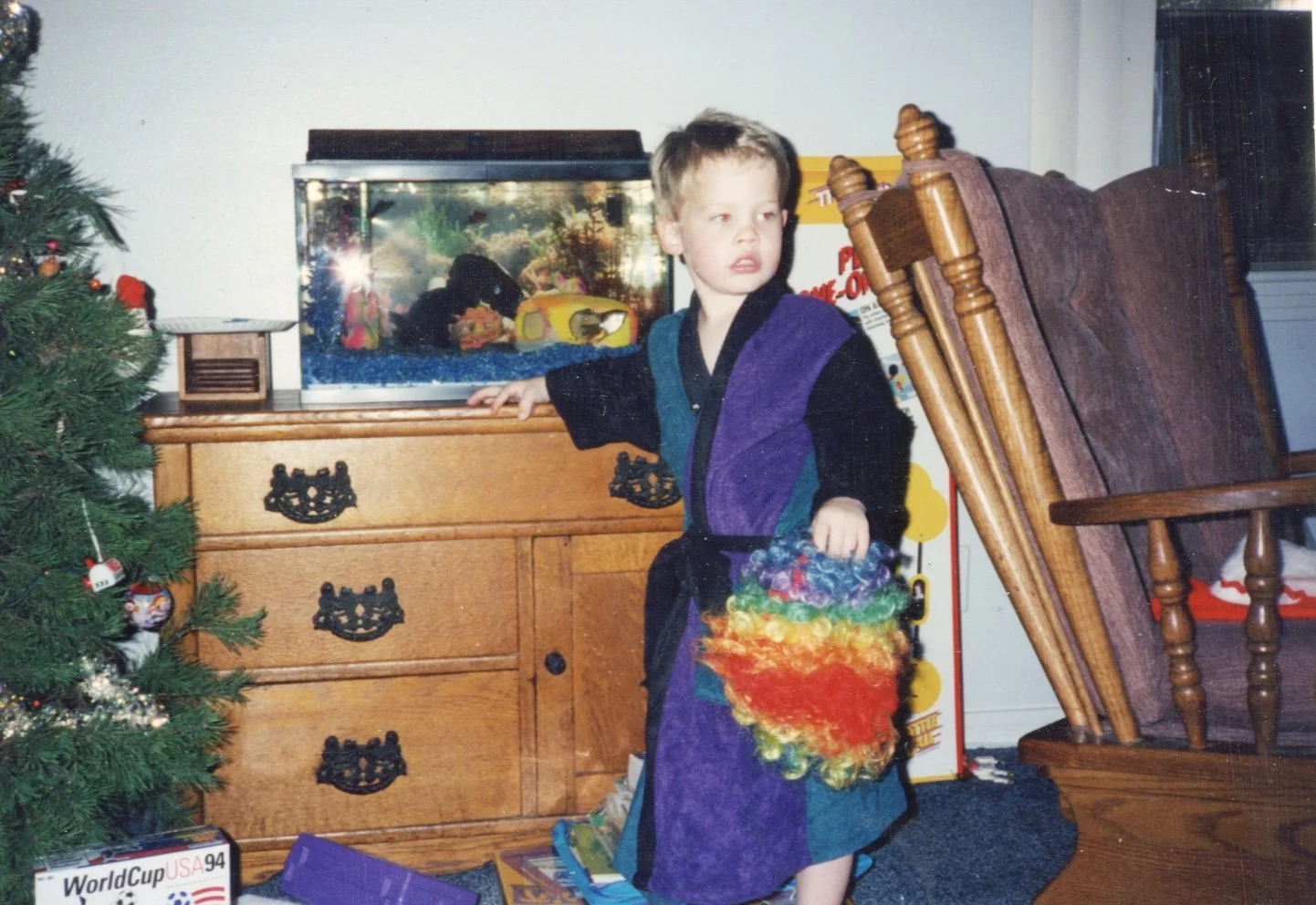 A young boy dressed in a colorful puppet costume standing next to a wooden dresser with a fish tank on top, in a room decorated for Christmas with a Christmas tree and ornaments.