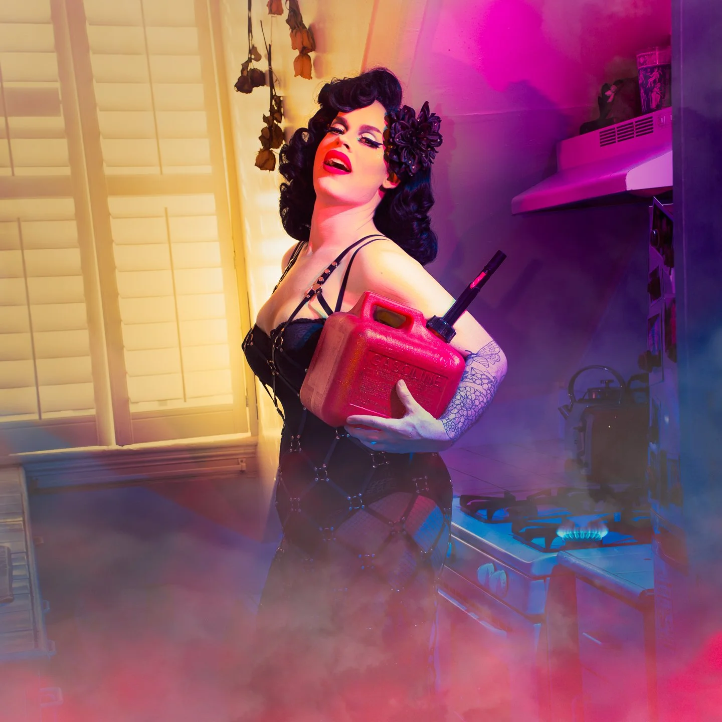 A woman with dark curly hair, dressed in black lingerie, holding a red gasoline can with a funnel, standing in a brightly colored kitchen with pink and blue lighting effects.