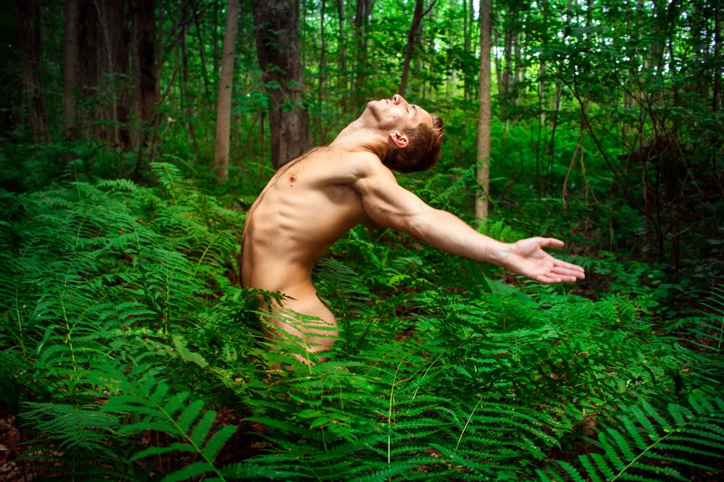A shirtless man with closed eyes and an expressive face stands with arms outstretched among lush green ferns and trees in a dense forest, suggesting a pose of freedom or connection with nature.