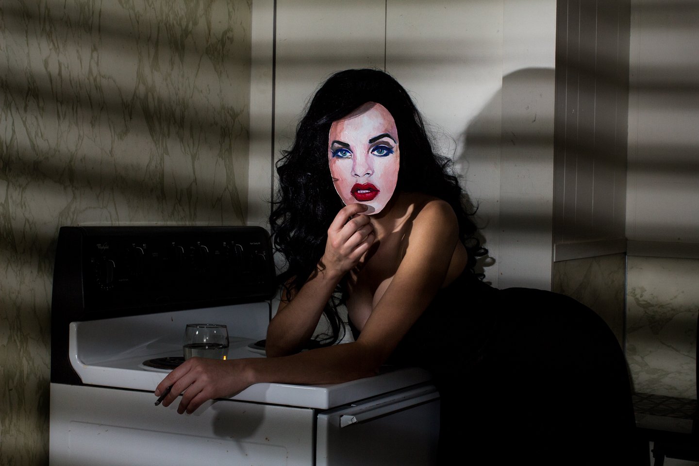 A woman with long black curly hair, wearing a black dress, holds a mask with a painted female face in front of her face. She is leaning on a stovetop with a glass of water in front of her. The background consists of marble and a kitchen wall.