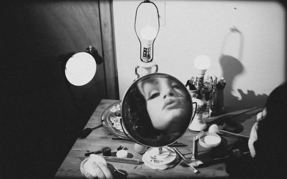 A mirror on a cluttered wooden table reflecting a woman's face with makeup, surrounded by small makeup containers, brushes, and a lamp. The background shows a door and a wall with shadows.