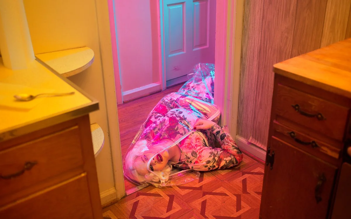 A person wearing a floral dress lying on the floor just inside a doorway, with a clear plastic umbrella covering them, illuminated by pink and blue lighting in a kitchen with wooden cabinets.
