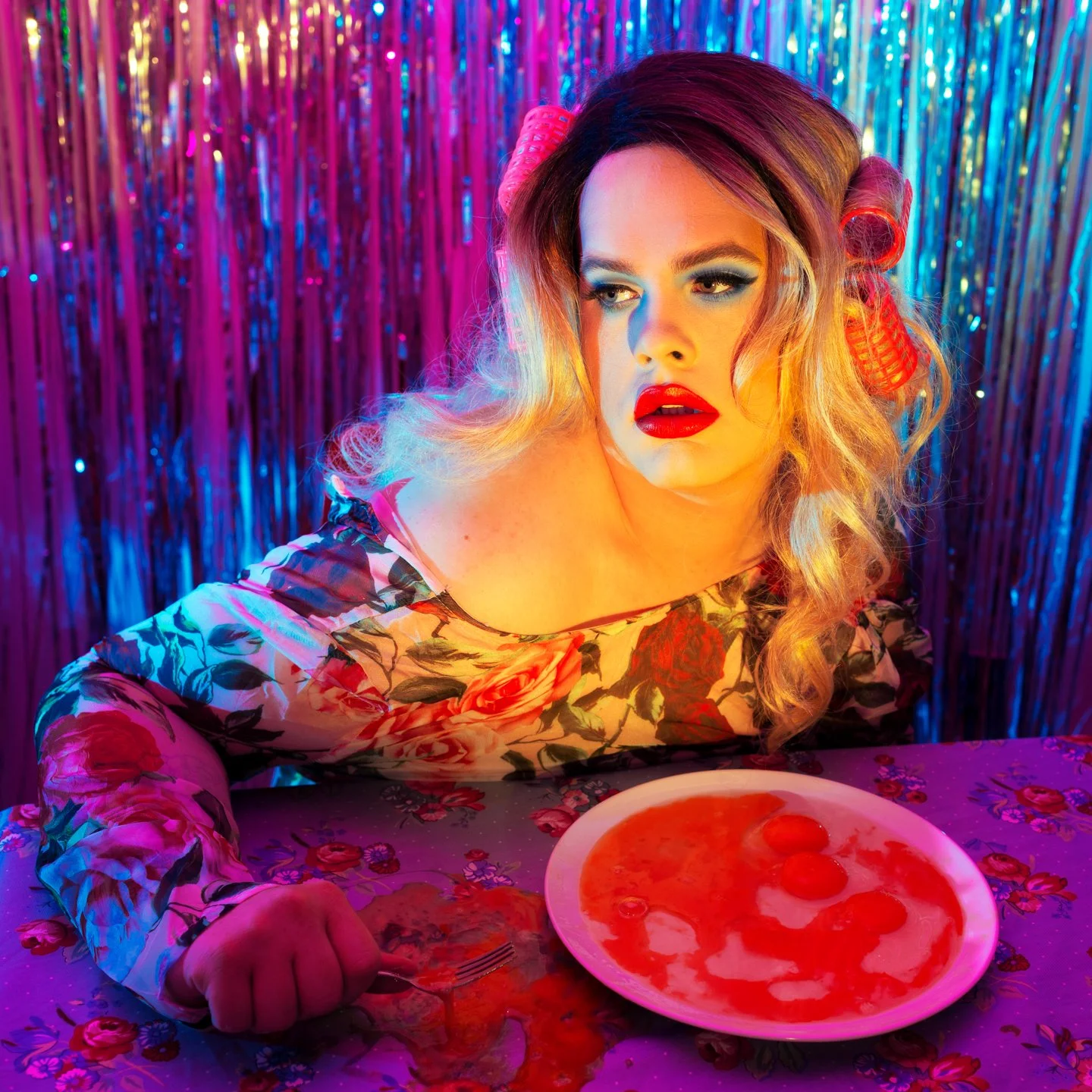 Person with colorful hair and bold makeup sitting at a table with a spilled plate of pizza, colorful background, and neon lighting.