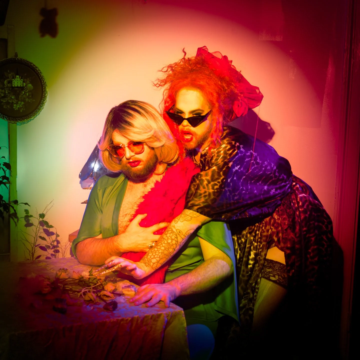 Two individuals with exaggerated makeup and colorful clothing posing indoors, with one wearing sunglasses and the other with heavy eye makeup and bright red hair, in a playful, avant-garde style.