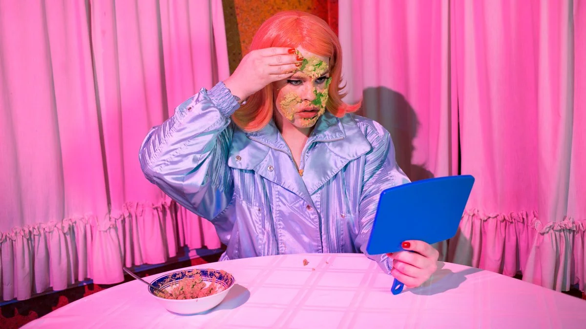 Woman with peach-colored hair, wearing a shiny gray jacket, looking into a mirror with a surprised expression, with green and yellow food smeared on her face, sitting at a table with a bowl of food, pink curtains in the background.