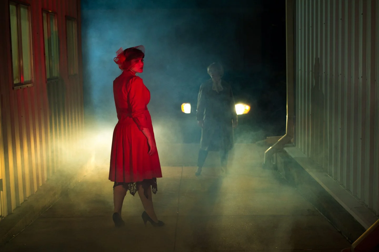 A woman in a red dress and hat standing in a foggy, dimly lit outdoor area at night, with a man in the background near car headlights, and industrial-style metal-sided buildings on either side.