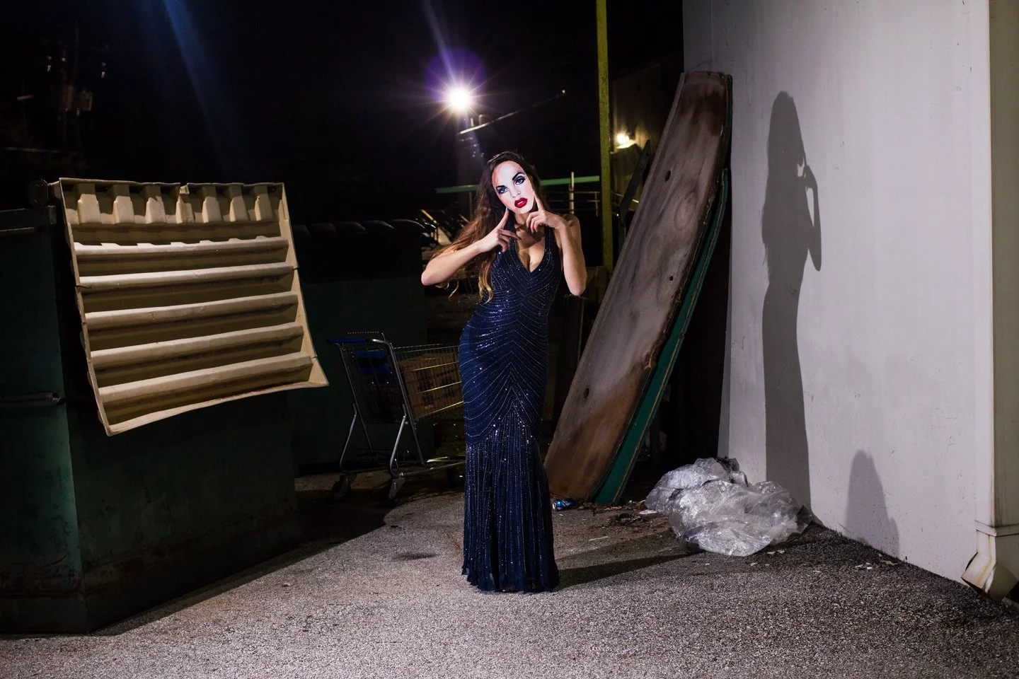 A woman in a sparkling navy blue evening gown posing at night outdoors, with a spotlight casting her shadow on a white wall.