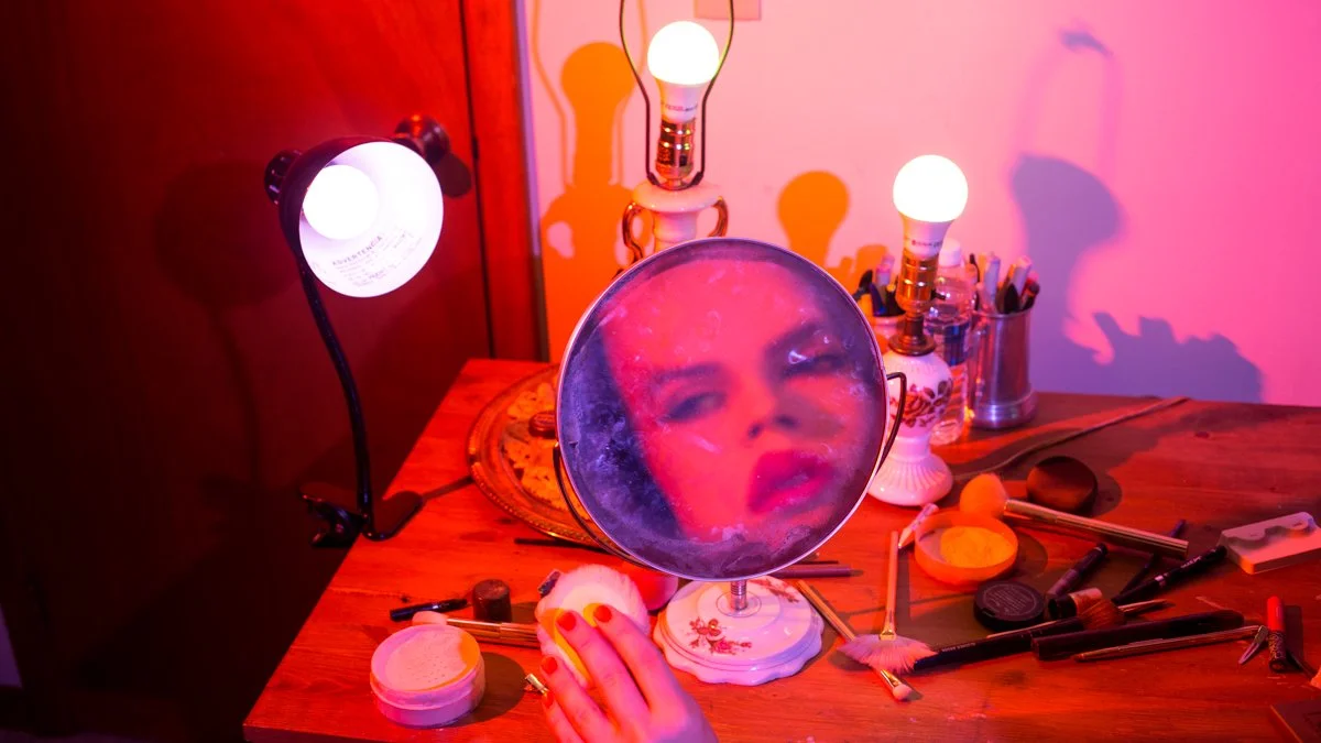 A person applying makeup at a wooden table cluttered with makeup tools, with a mirror reflecting their face and three light bulbs illuminating the scene.