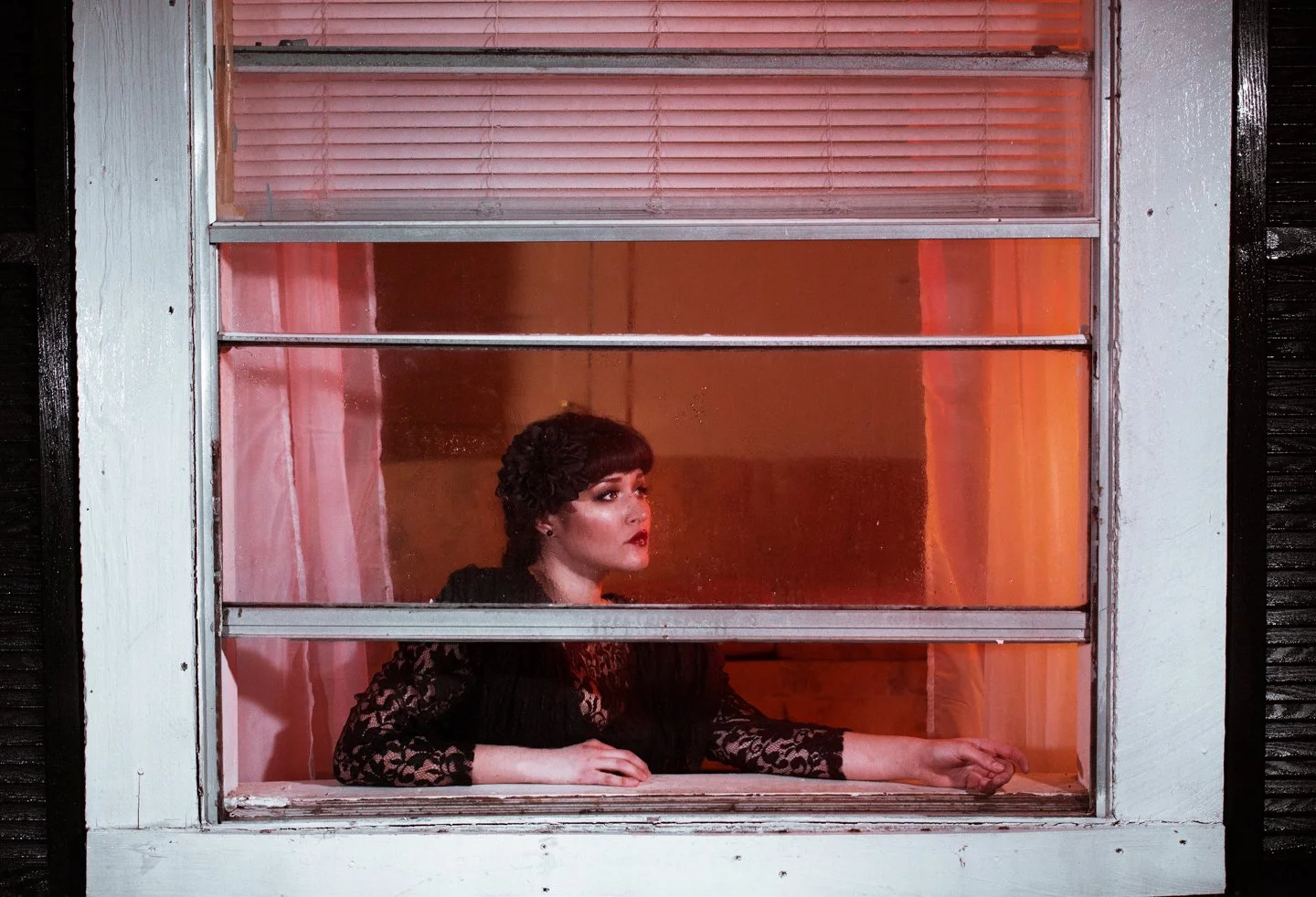Woman with dark hair and red lipstick looking out of a window with pink curtains behind her, wearing a black lace top, inside a building at night.