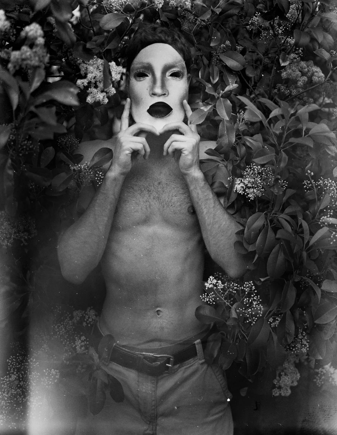 A person with a mask of a face, lying on a bed of leaves and flowers, with their hands holding their face mask and chest.