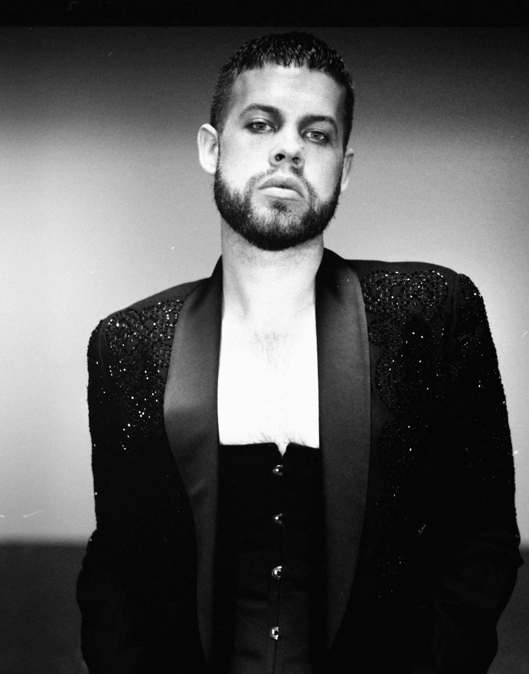 A black and white portrait of a man with short hair, a beard, wearing a jacket with sequined shoulder accents and a low-cut shirt underneath.