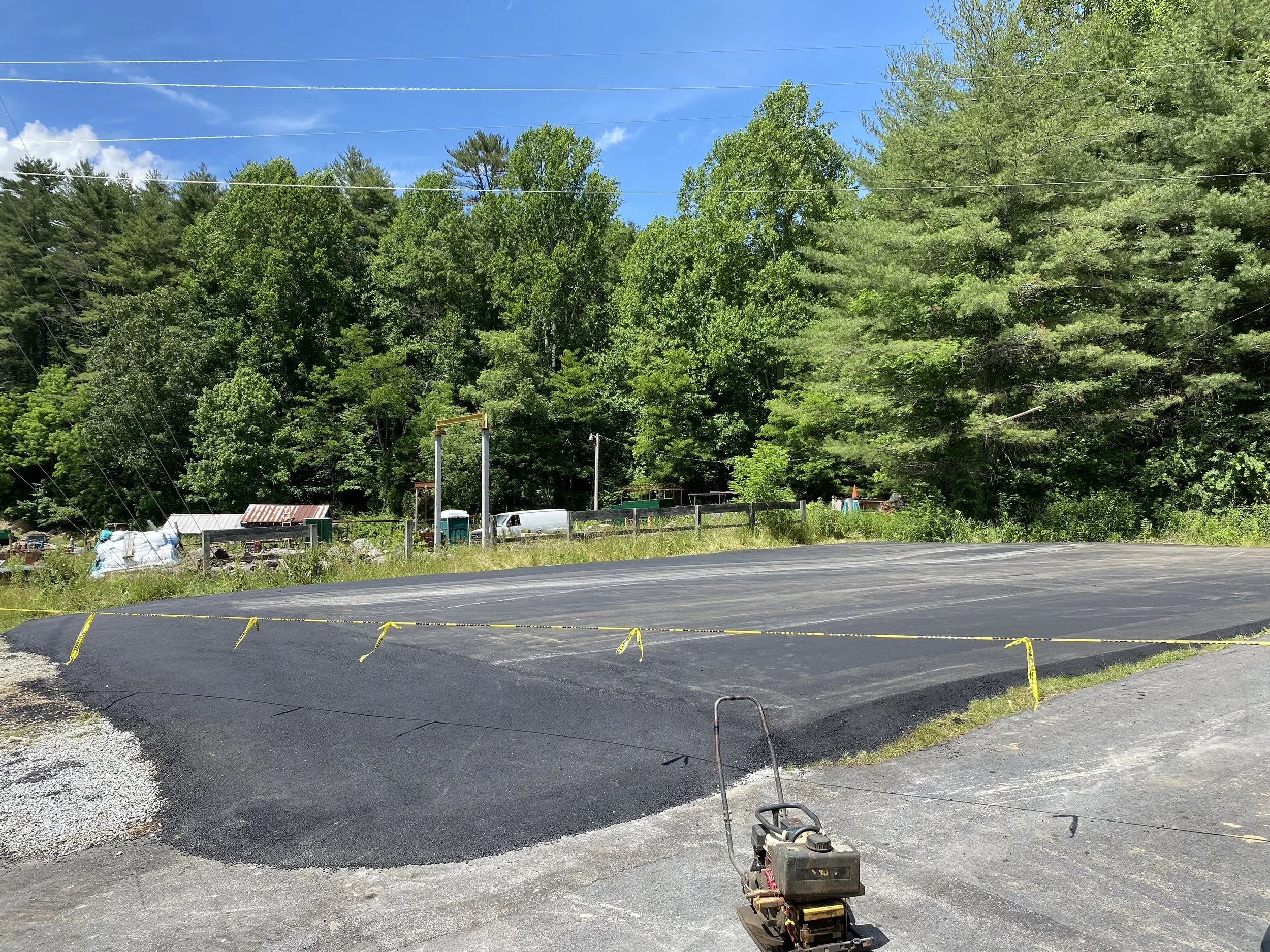 A freshly paved asphalt driveway under construction with a small plate compactor in the foreground, surrounded by green trees and blue sky.