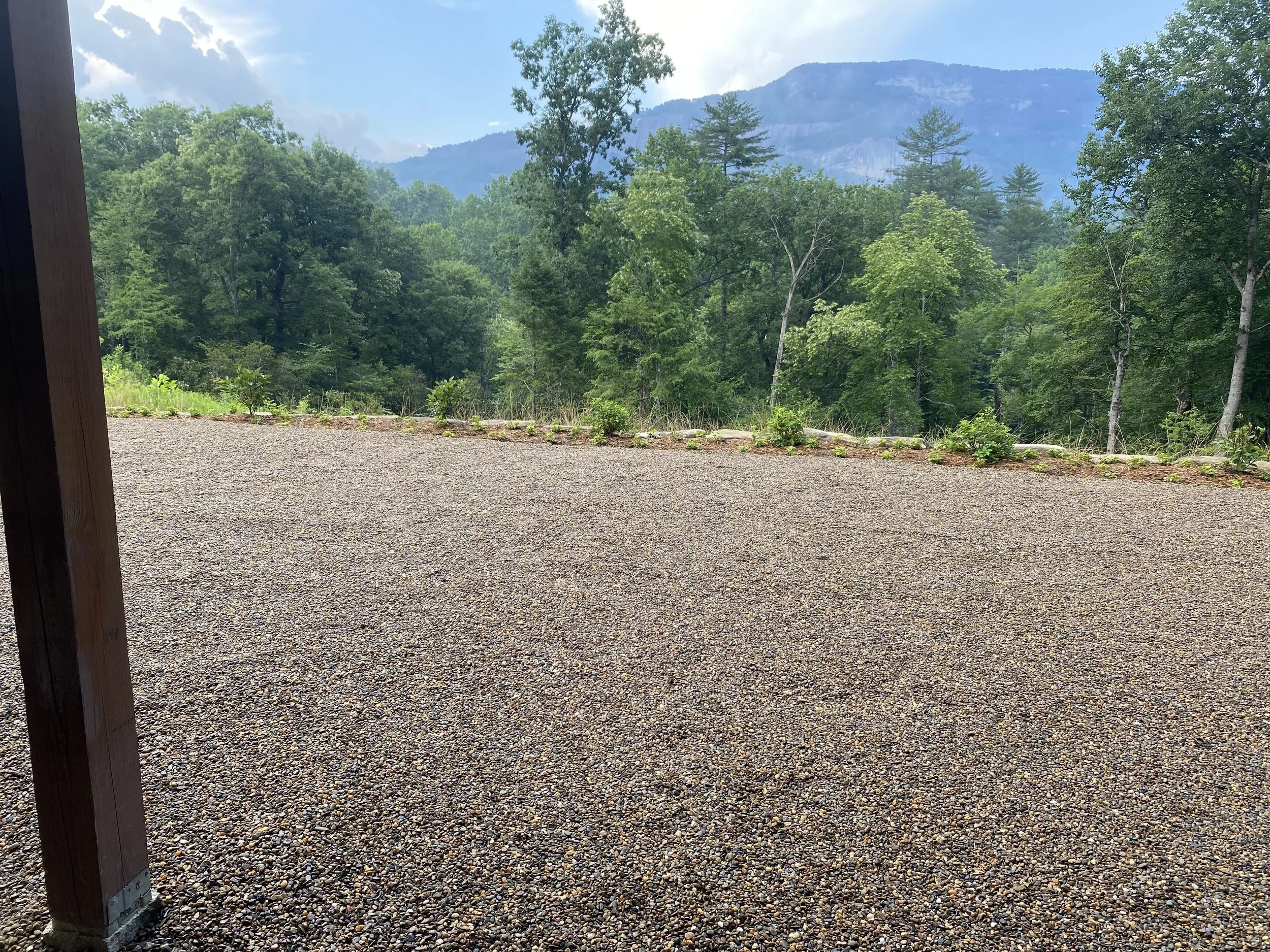 View of a forested mountain landscape with trees and mountains in the background, seen from a gravel area with a wooden post on the left.
