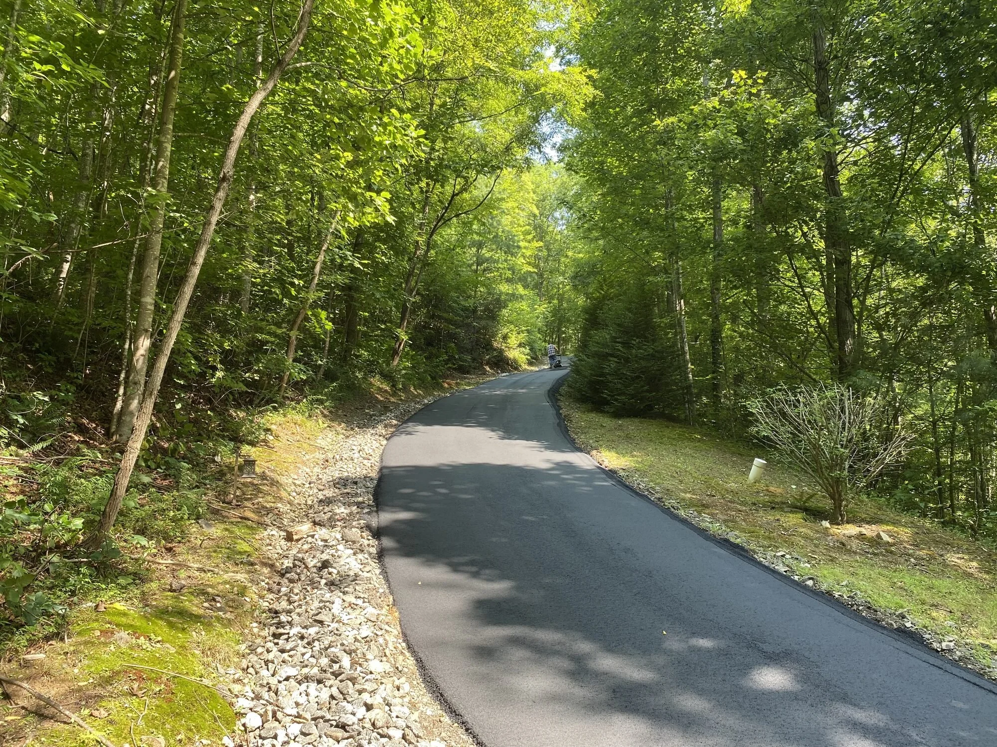Smooth finished asphalt driveway winding through a wooded lot with a fresh gravel shoulder in Whittier, Jackson County NC — paved by Black Bear Paving