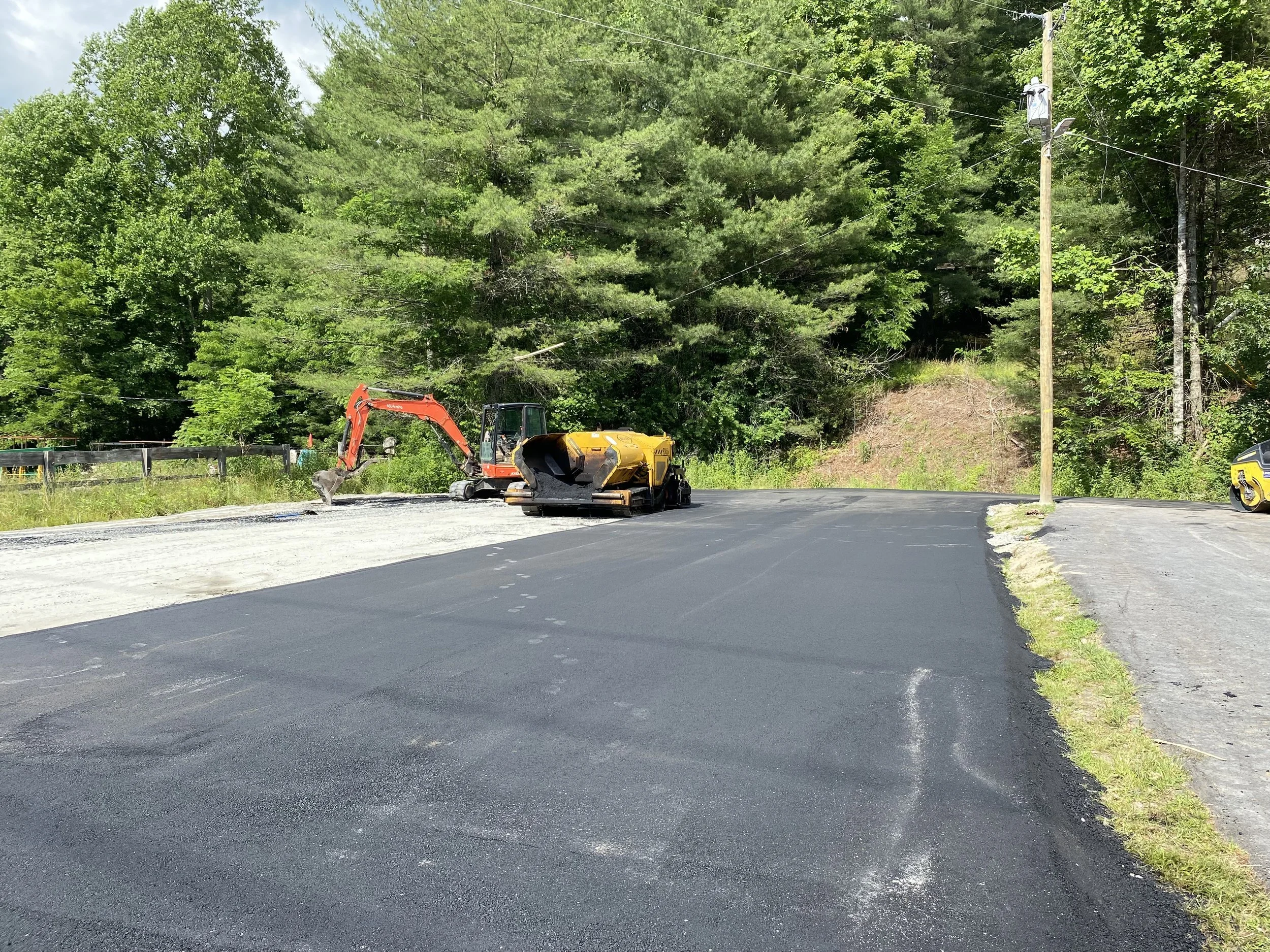 A construction site on a road with fresh black asphalt, a small excavator, and a roller compactor near the edge, surrounded by trees and utility poles.
