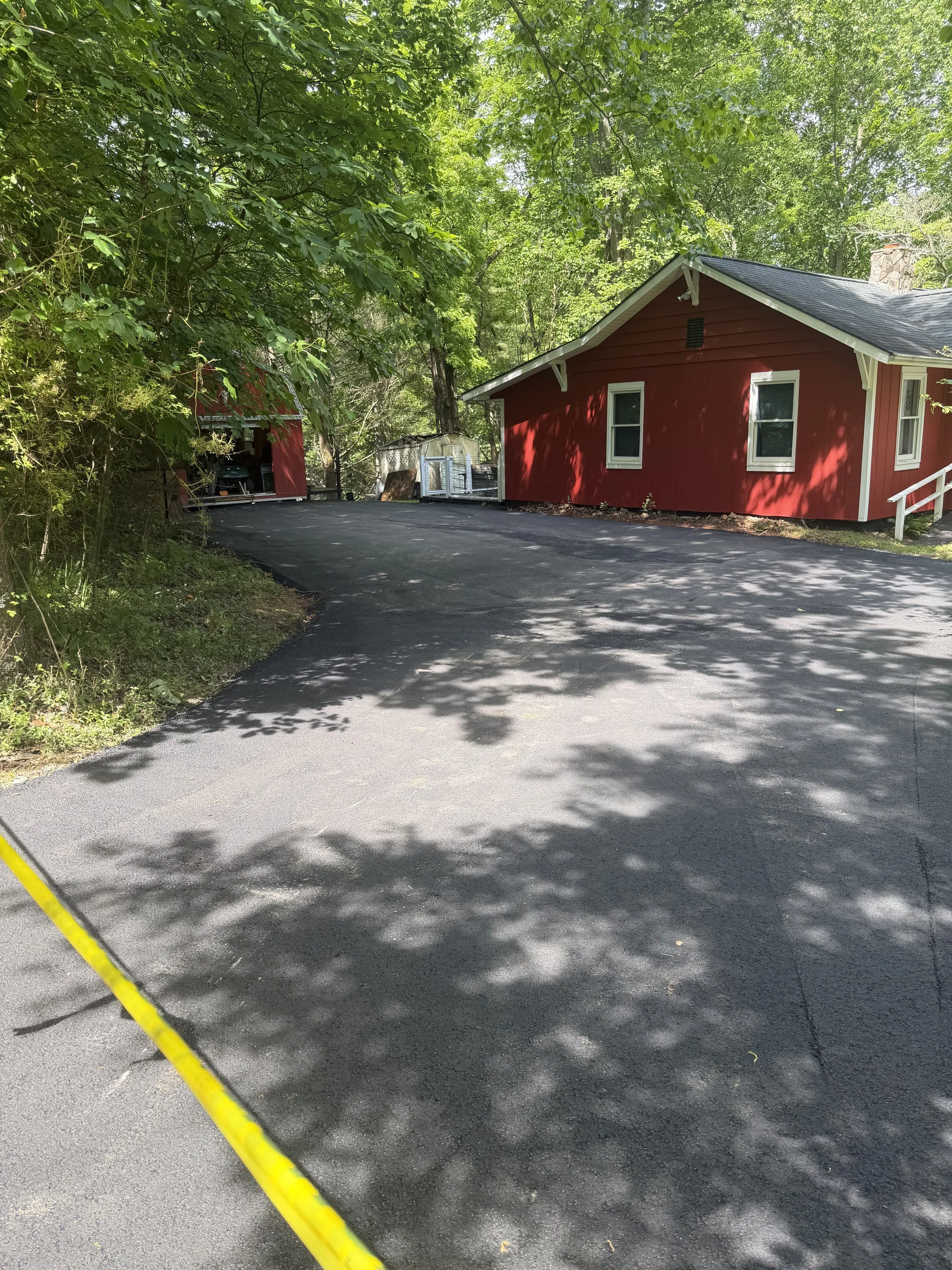 A paved driveway in a wooded area with shadows of trees, a red house with white trim, a small shed, and a trailer in the background.
