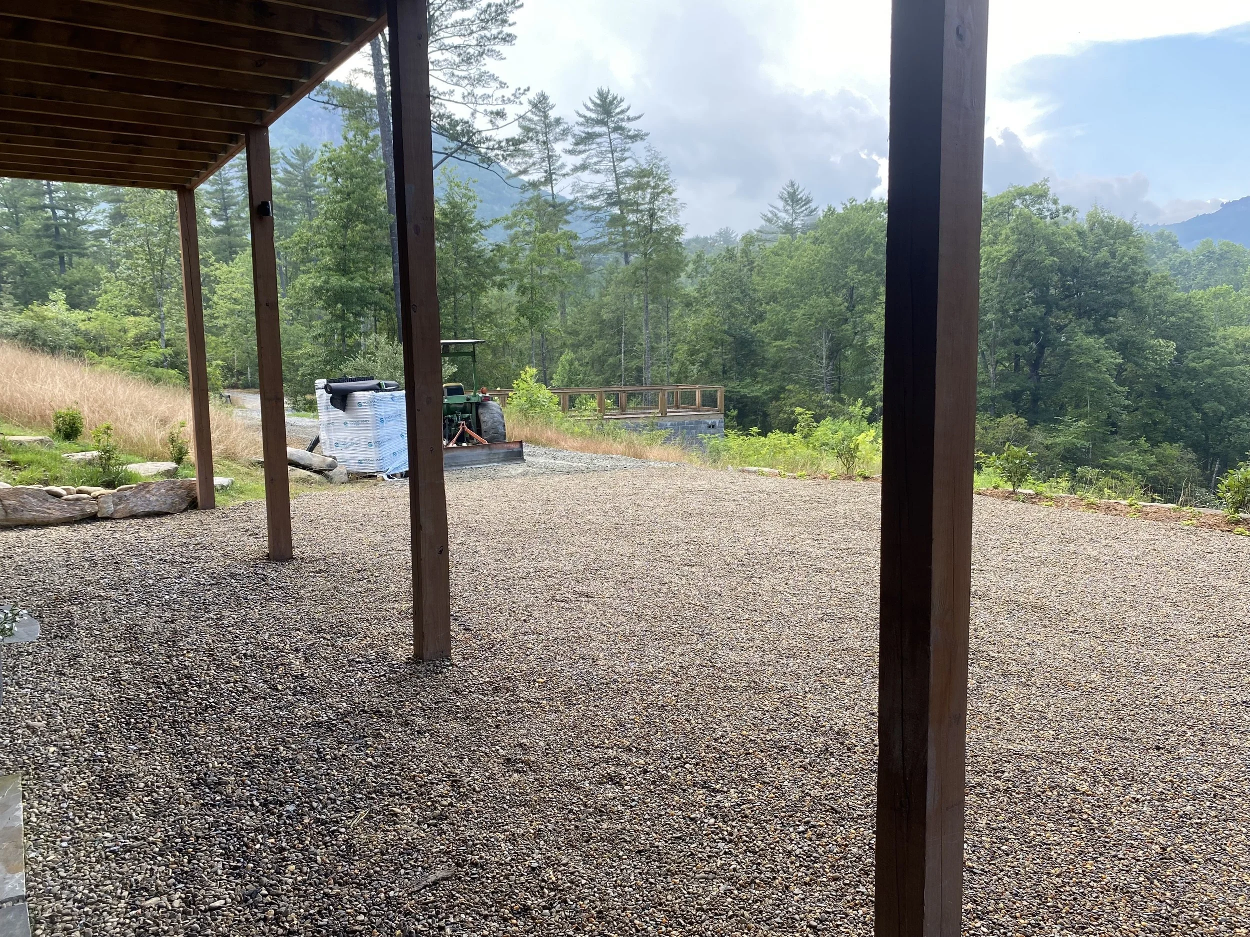 View from a porch showing a gravel ground, green trees, a tractor, and mountains in the distance under a partly cloudy sky.