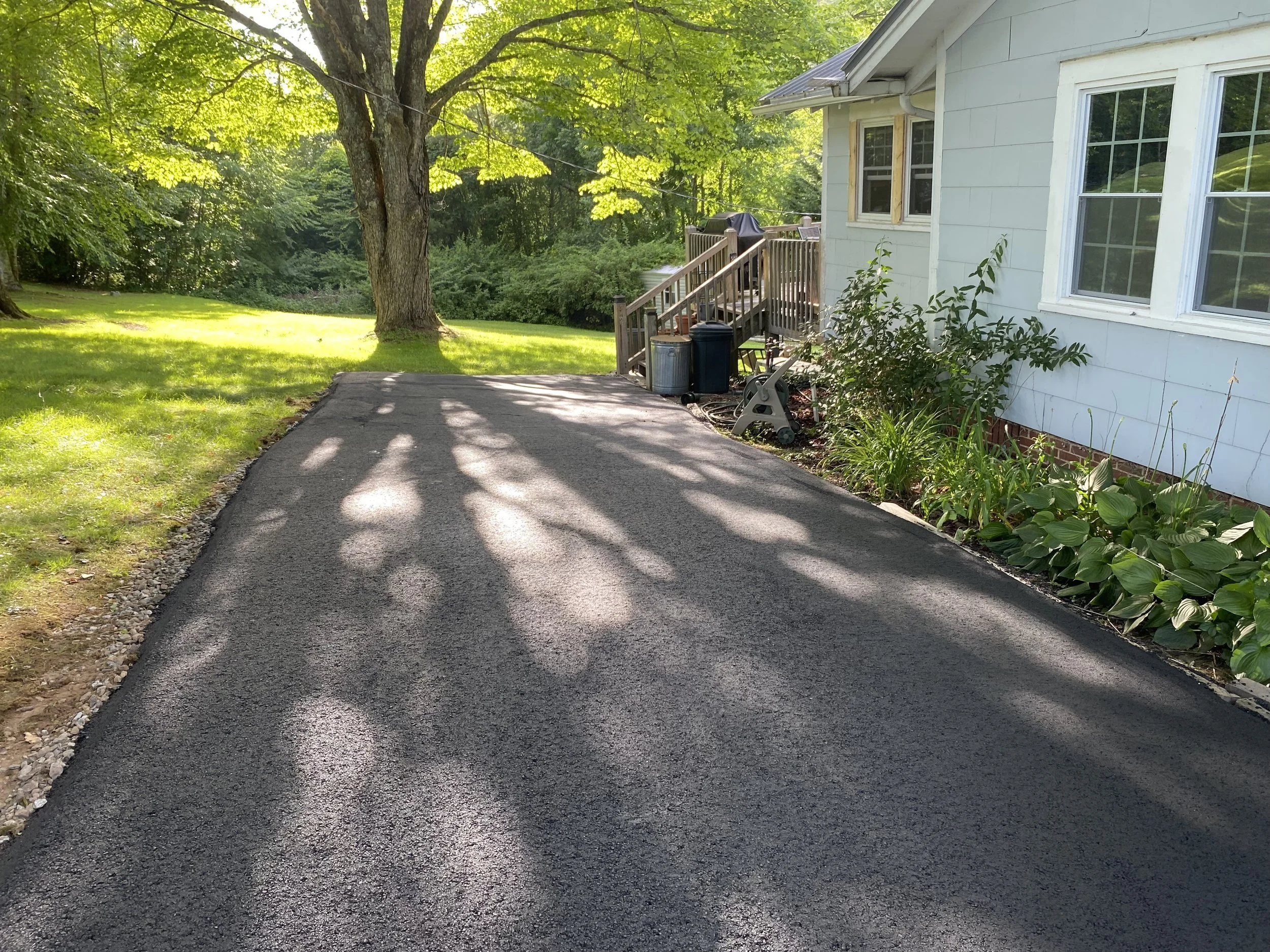A paved driveway next to a house with white siding and multiple windows. There are garden beds with green plants along the house and a grassy yard with a large tree casting shadows on the driveway. In the background, there are bushes and trees.