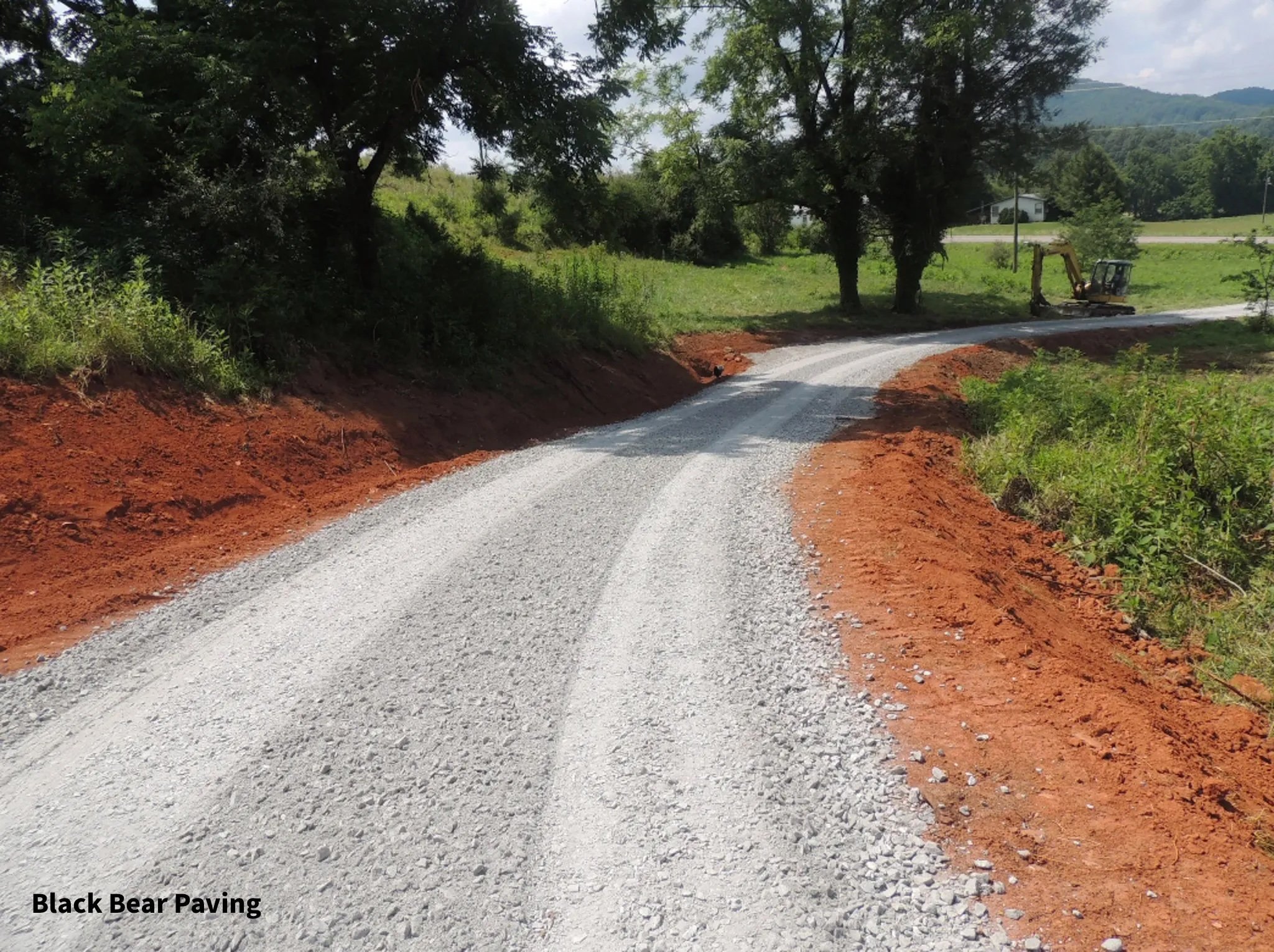 Gravel road with red dirt sides, surrounded by green trees and hills in the background, construction equipment on the side, under partly cloudy sky.