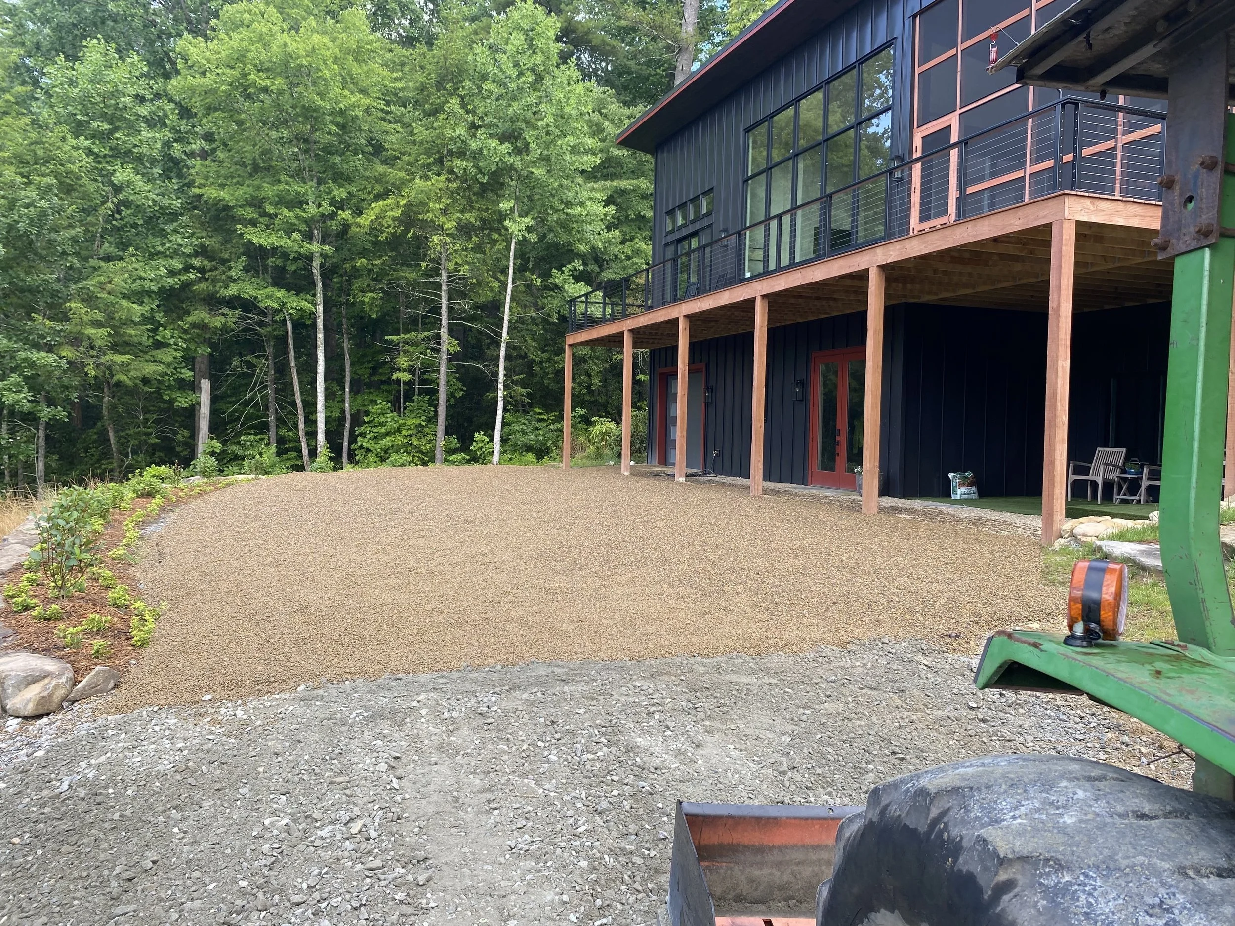 A house under construction with a black exterior and large glass windows, surrounded by green trees. A lot of gravel covers the ground in the foreground, with some construction tools and materials nearby.