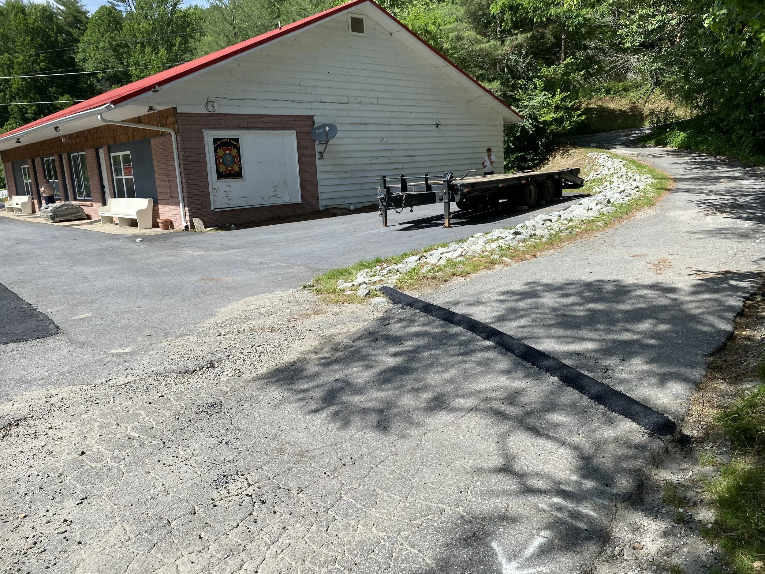 A small building with a red roof, white wood siding, and brick accents. A sign indicating it is a fire station, alongside parked benches and a black flatbed trailer. Two people are standing near the building, surrounded by trees and greenery, with a 