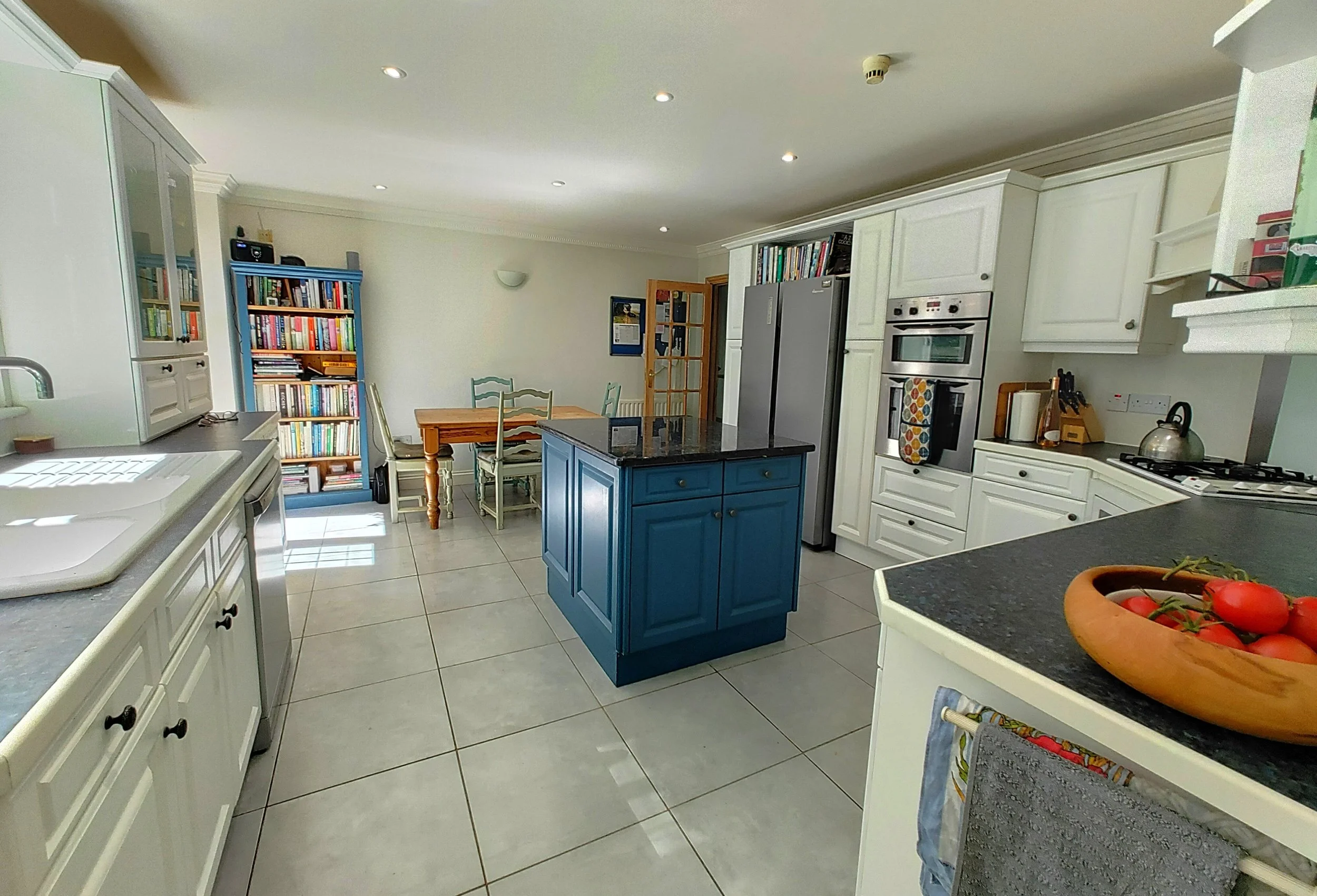 A bright kitchen with white cabinets, a blue kitchen island, a stainless steel refrigerator, a wooden dining table with chairs, a bookshelf with books, and a bowl of tomatoes on the counter.