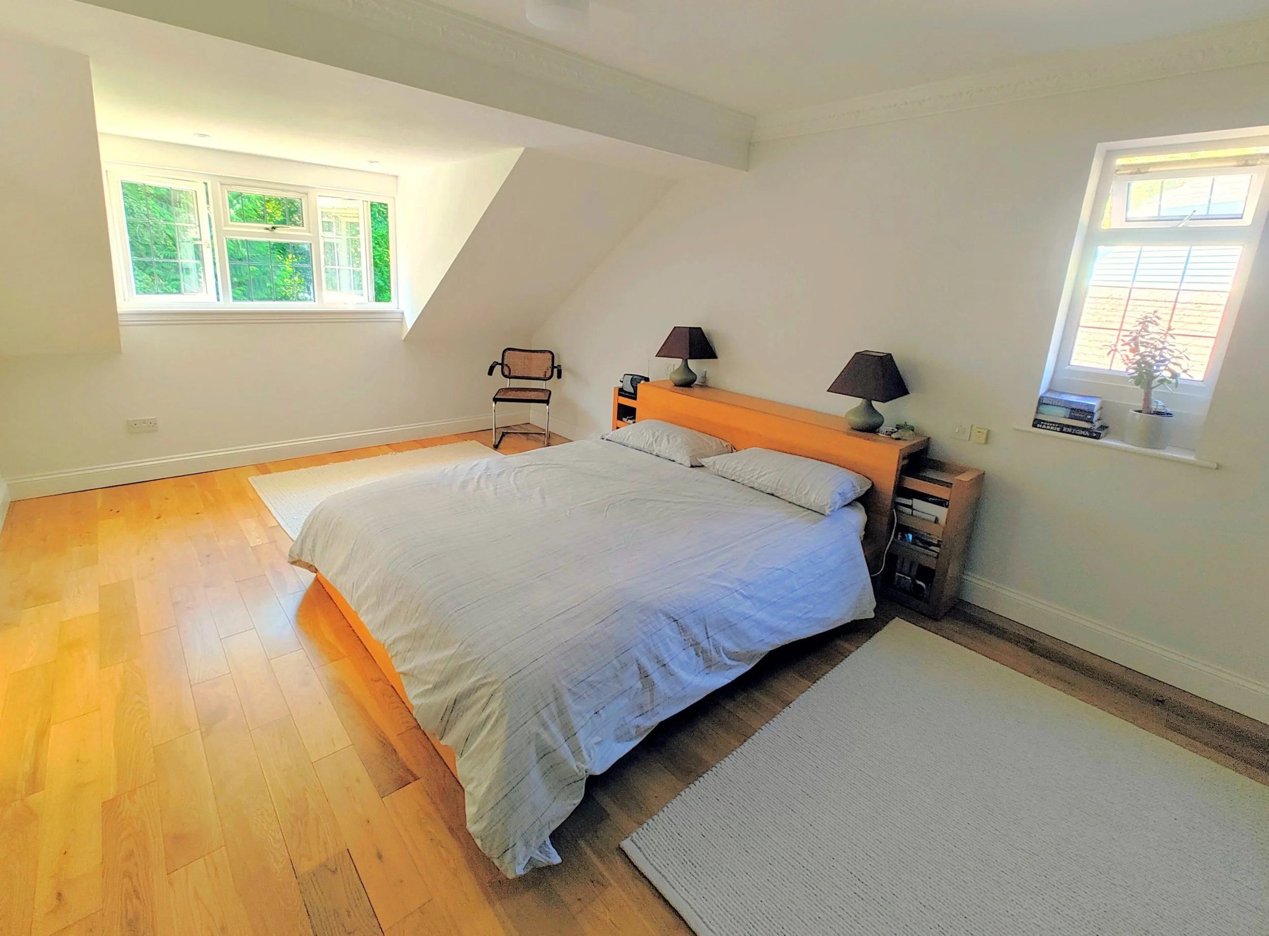 Bedroom with a bed, nightstands with lamps, a chair, large windows showing greenery, hardwood flooring, and a small plant on the windowsill.