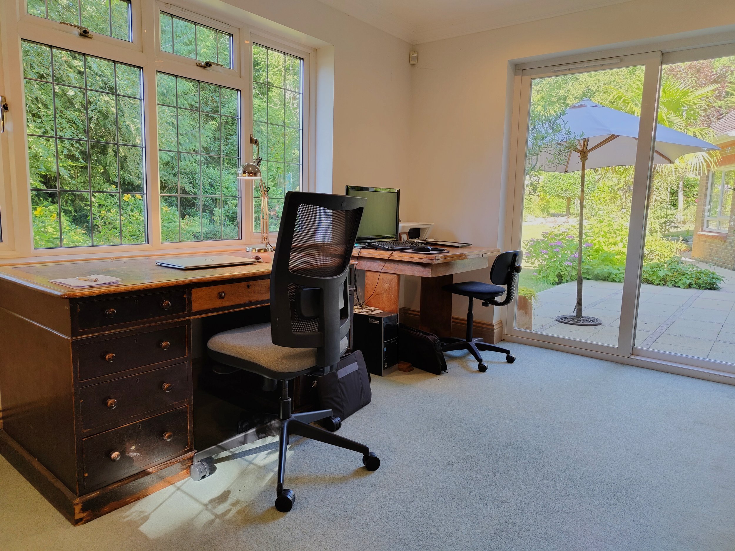 Home office with wooden desk, office chair, computer, and large windows showing outdoor plants and patio with umbrella.
