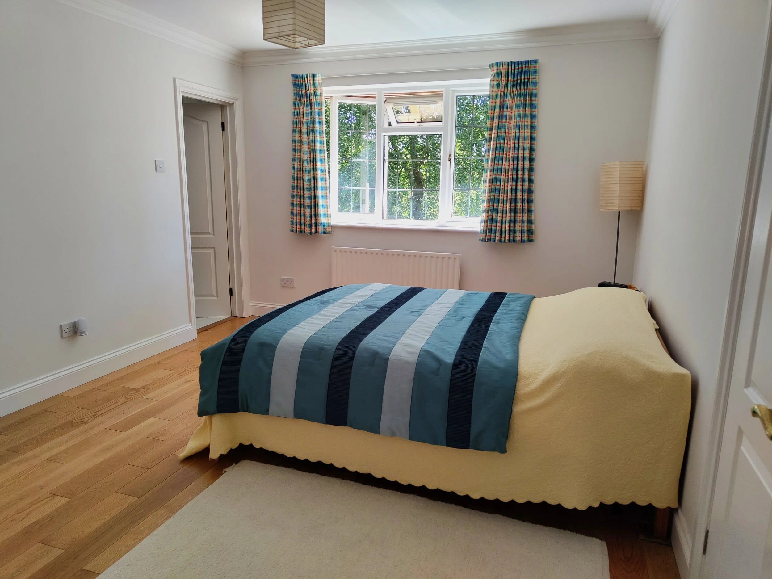 A bedroom features a window with checkered curtains, a bed with a striped quilt, a tall floor lamp, and hardwood floors.